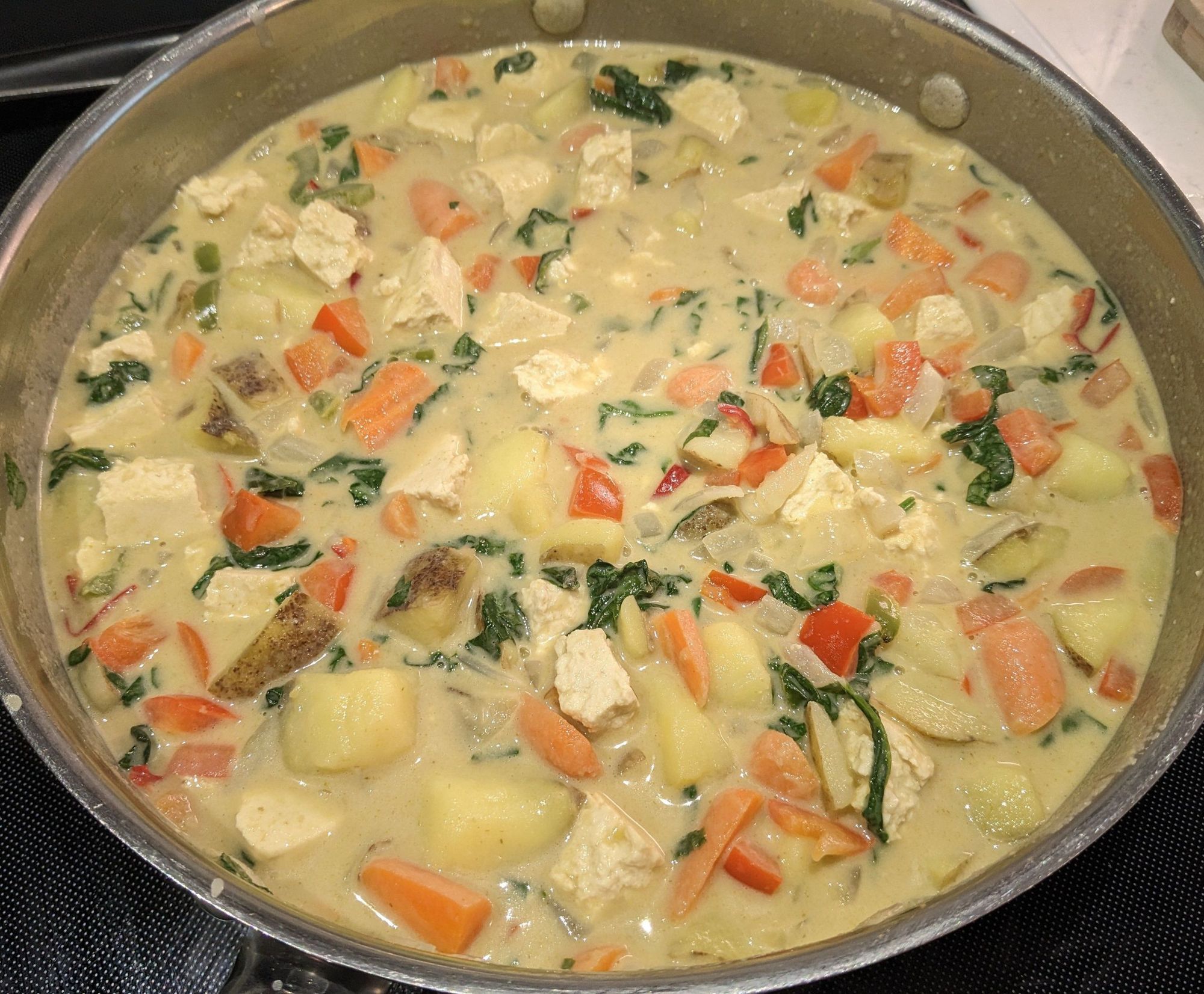 A cooked Thai-style Green vegetable curry in a large silver skillet with carrots, spinach, Chijimisai, tofu, potatoes, hot and sweet peppers. 