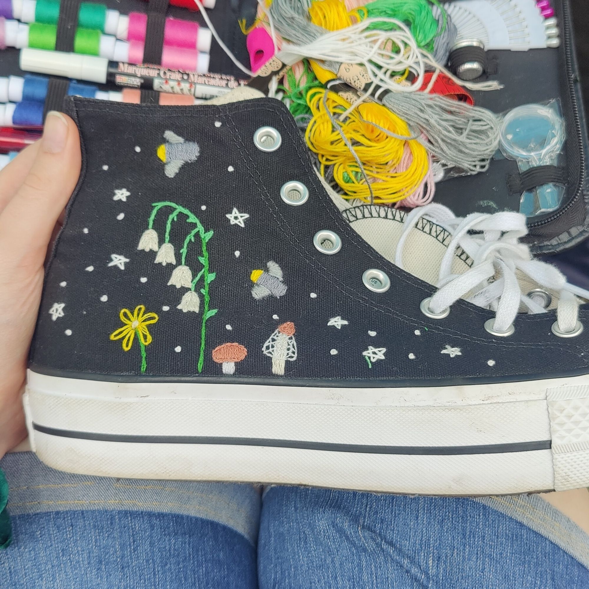 Embroidered converse with flowers mushrooms and fireflys with stars and dots as stars. The shoe is sitting on a lap with a sewing kit in the back