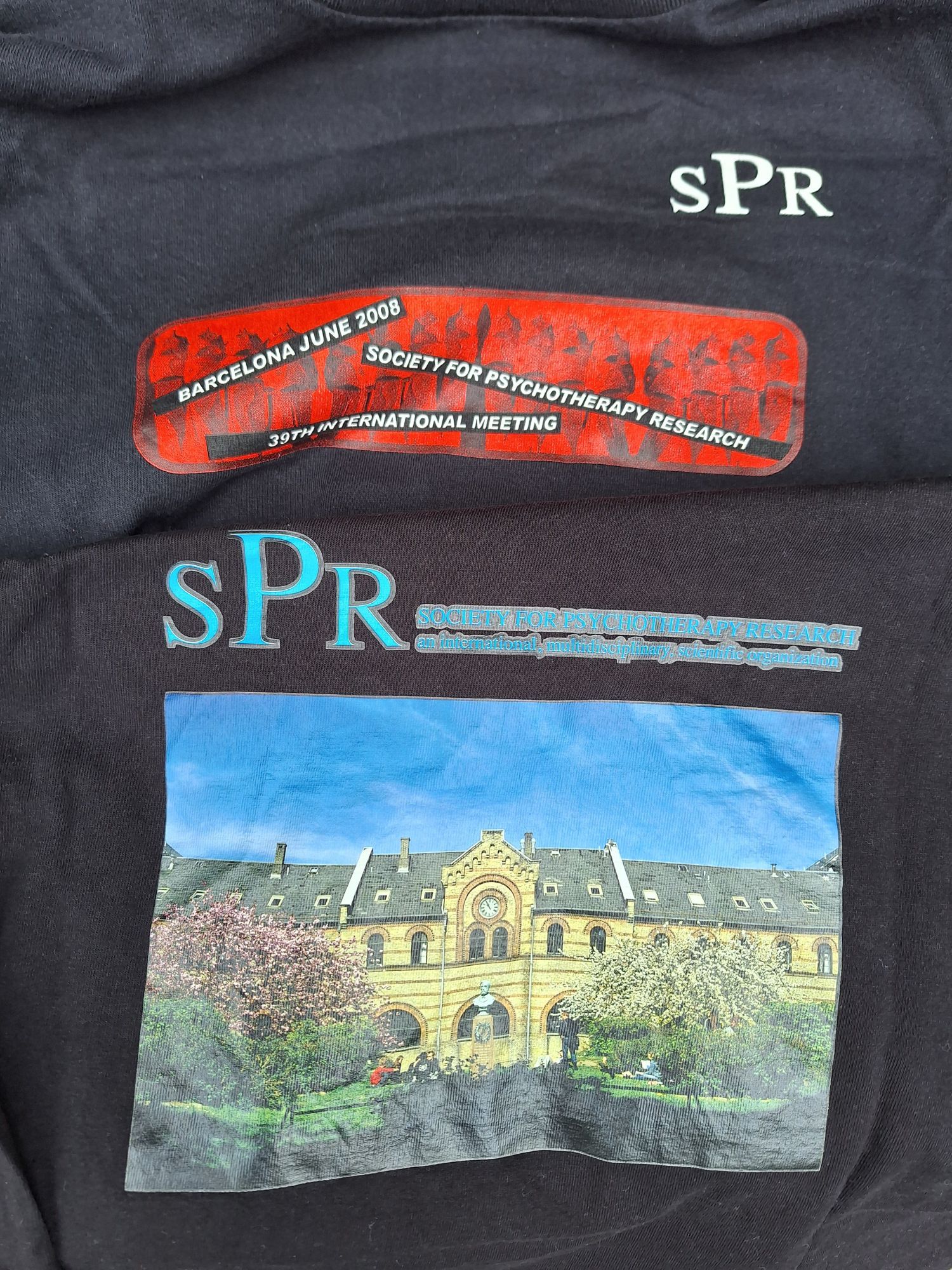 Picture of two black T-Shirts with prints on it, the top in red and black for the 2008 SPR meeting in Barcelona with stylised Gaudi elements; the bottom showing a picture of a building of Copenhagen University where the 2014 meeting was held.