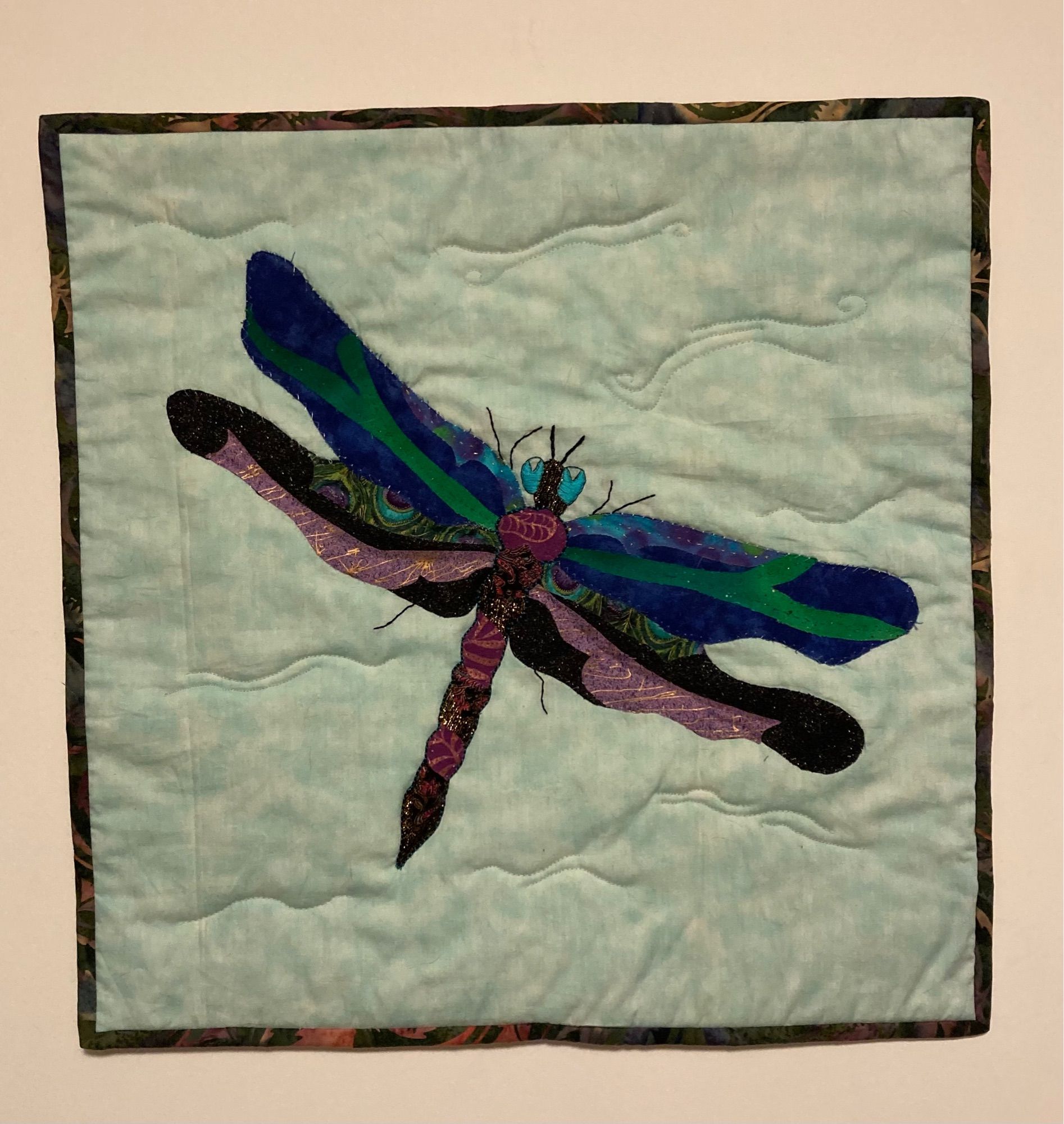 A quilted dragonfly on a light blue background. Its front wings are shades of green and blue, while the back wings are purple and black. It has bright blue embroidered eyes.