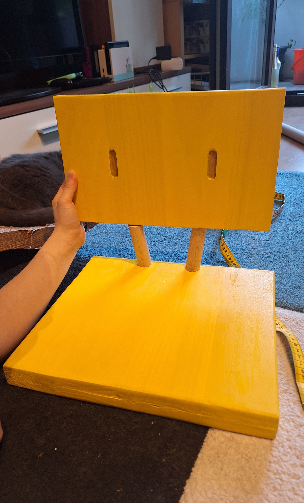 Yellow chair base on the carpet. The chair back has eyes and is supported by a hand.