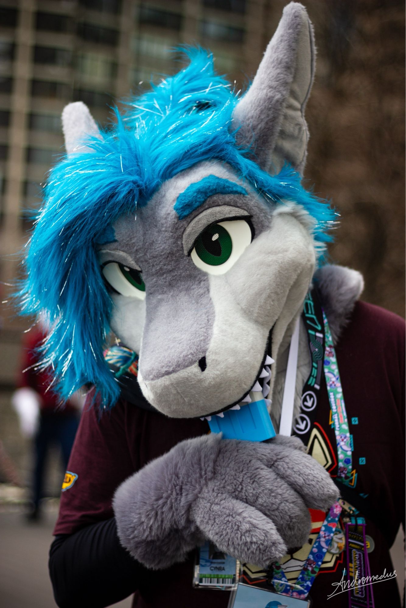 A shark fursuiter with a popsicle shaped fan in their mouth, looking cute towards the camera.