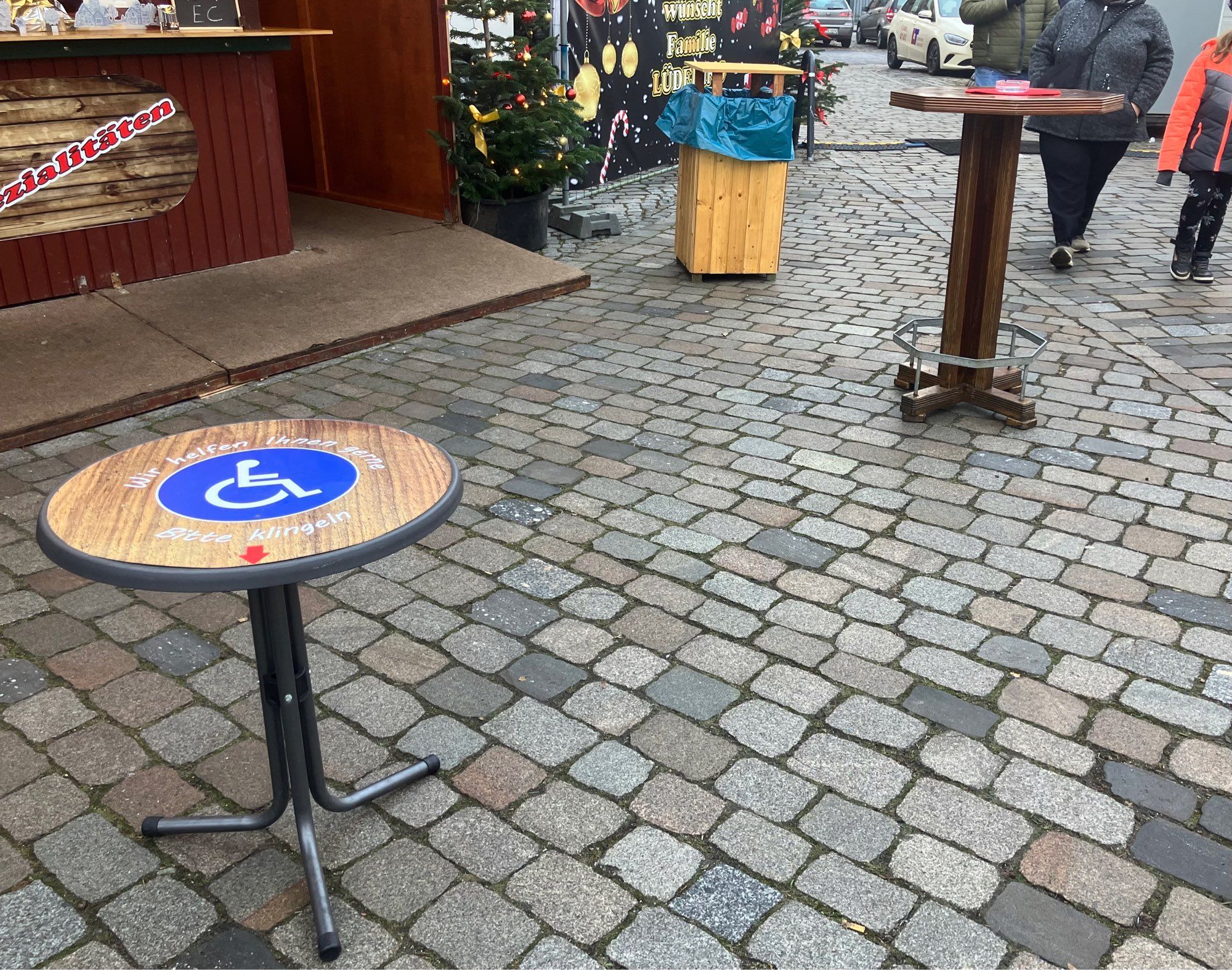 Gepflasterter Bereich vor einem Grillstand mit zwei Stehtischen, davon einer deutlich niedriger. Auf der Tischplatte ein großes Rolli-Symbol und die Aufschrift „Wir helfen Ihnen gerne“.