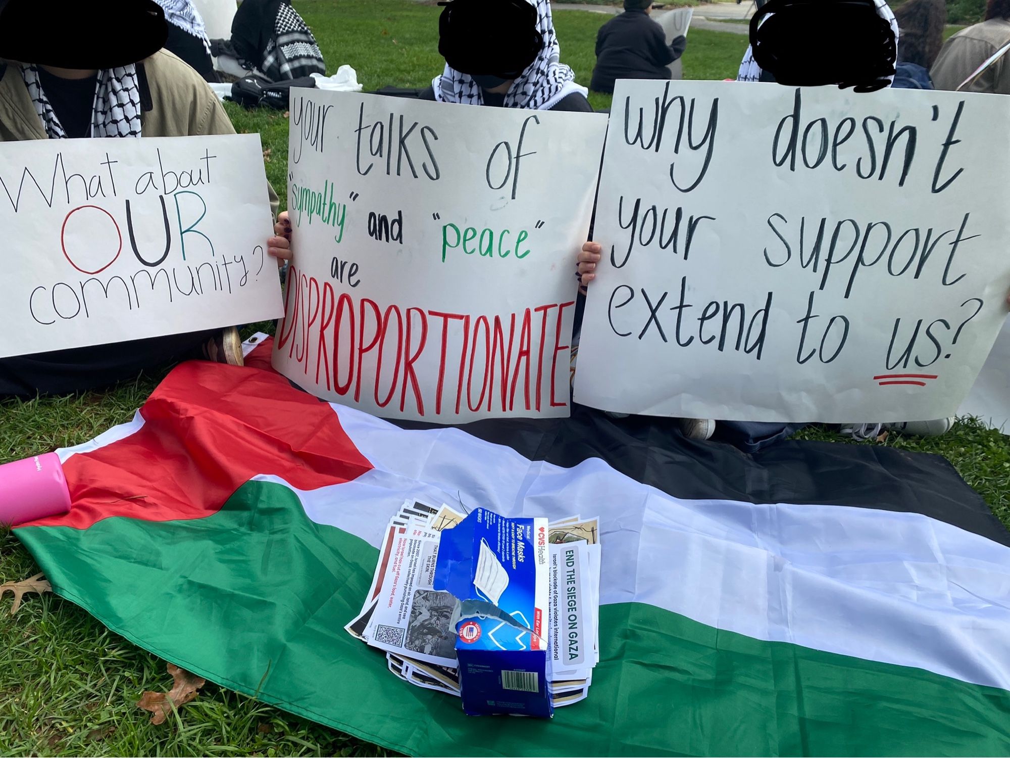 A circle of people sit on the lawn at American university, closer up are three people holding sings that say “what about our community” “your talks of sympathy and peace are disproportionate” and “why doesn’t your support extend to us?” In front of them is a spread out Palestinian flag and a pile of fliers and box of masks.