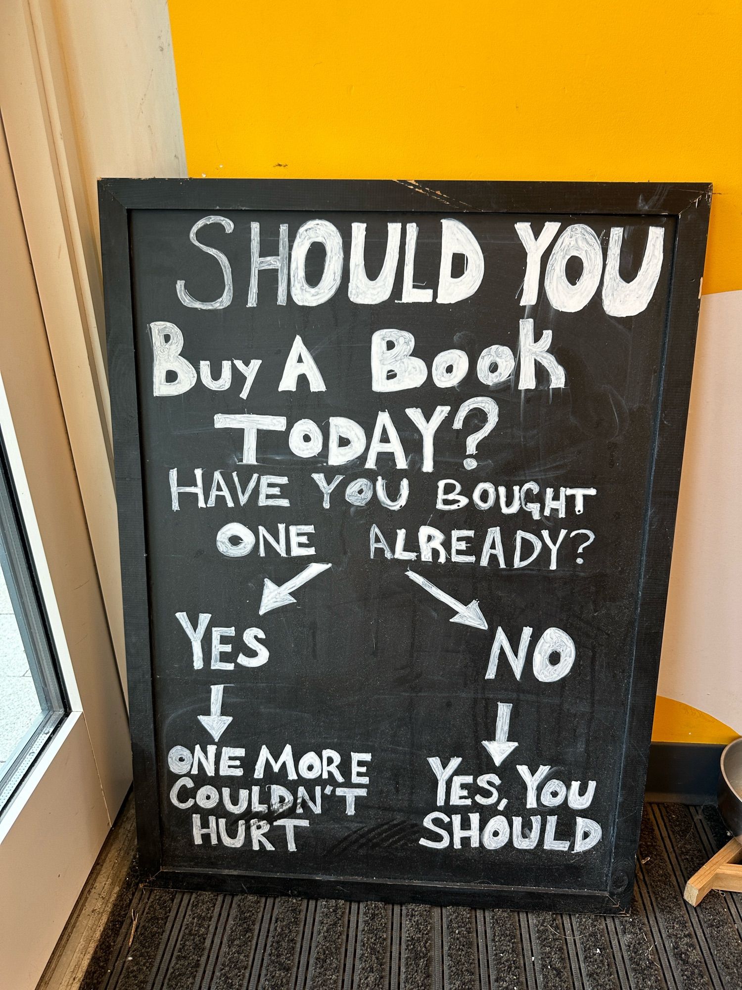 Handwritten board sign reading “Should you buy a book today?” with a flow chart below. It goes: “Have you bought one already? Yes -> one more couldn’t hurt and No -> yes, you should