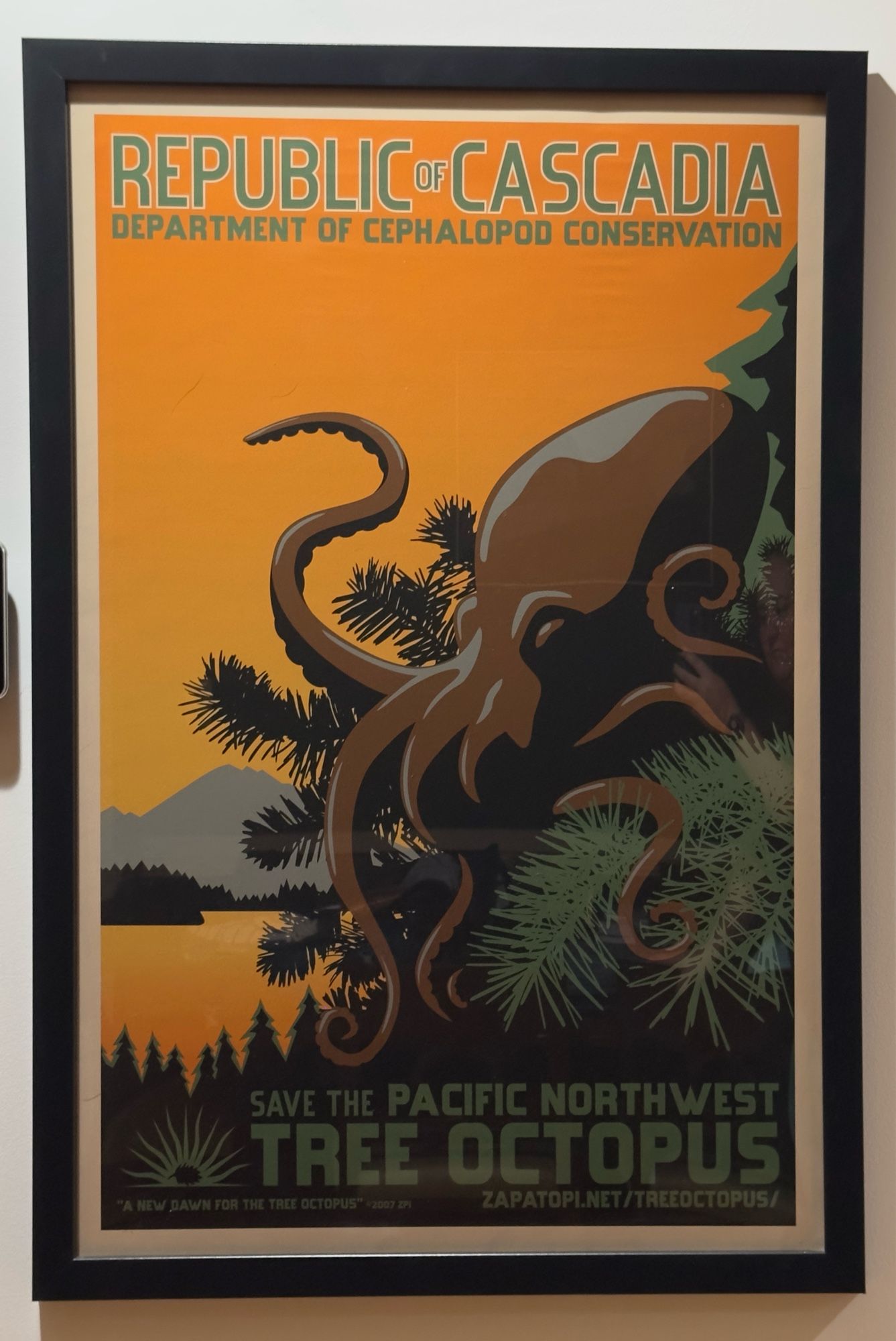 Poster for the Republic of Cascadia Department of Cephalopod Conservation’s campaign to save the Pacific Northwest Tree Octopus. On the poster, a brown and gray octopus perches amidst the branches of a pine tree, in front of a stereotypical Pacific Northwest lake sunset