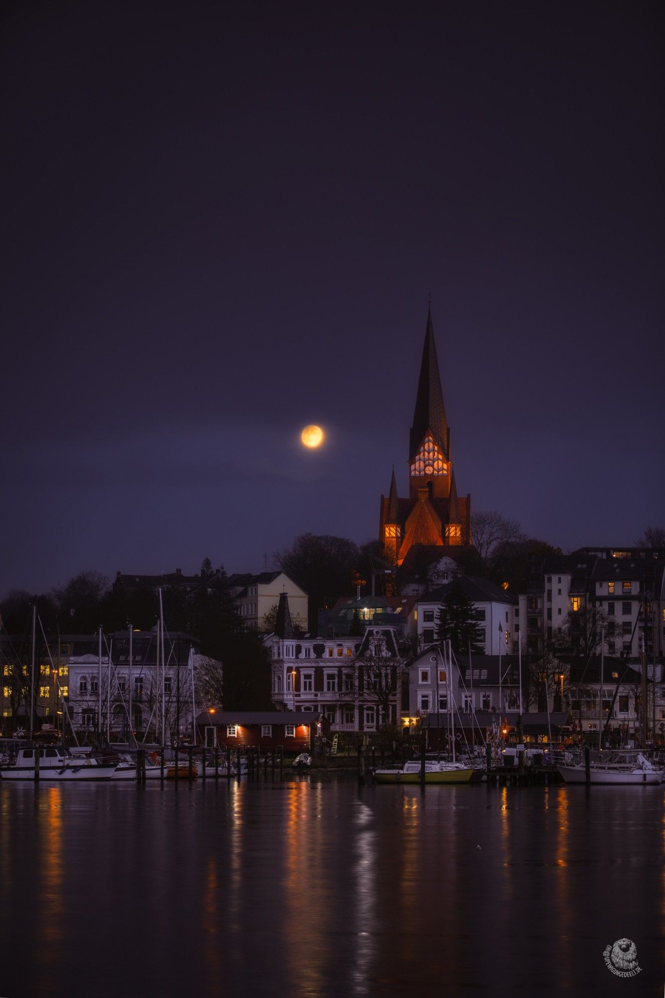 Flensburg, Blick aufs Ostufer in der Dämmerung. Es ist dunkel, leicht violettfarben, der Turm der St. Jürgen Kirche ist zu sehen, von unten angestrahlt, daneben ein Vollmond. Die wenigen Lichter spiegeln sich in der Förde.