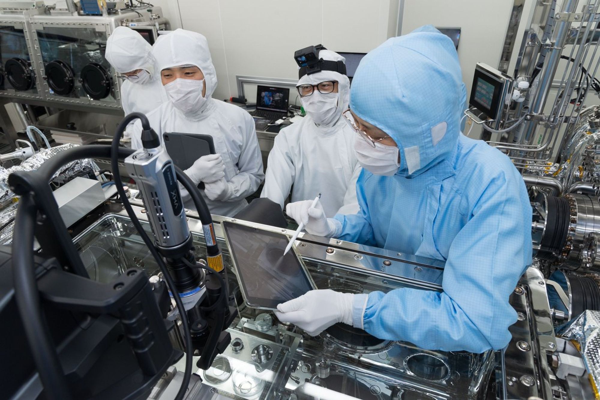 Photograph from inside the JAXA cleanrooms. Four people in PPE bunny suits are standing amongst the shiny metal equipment. One is tapping on a tablet with an electronic pencil. Another person is wearing a head camera.