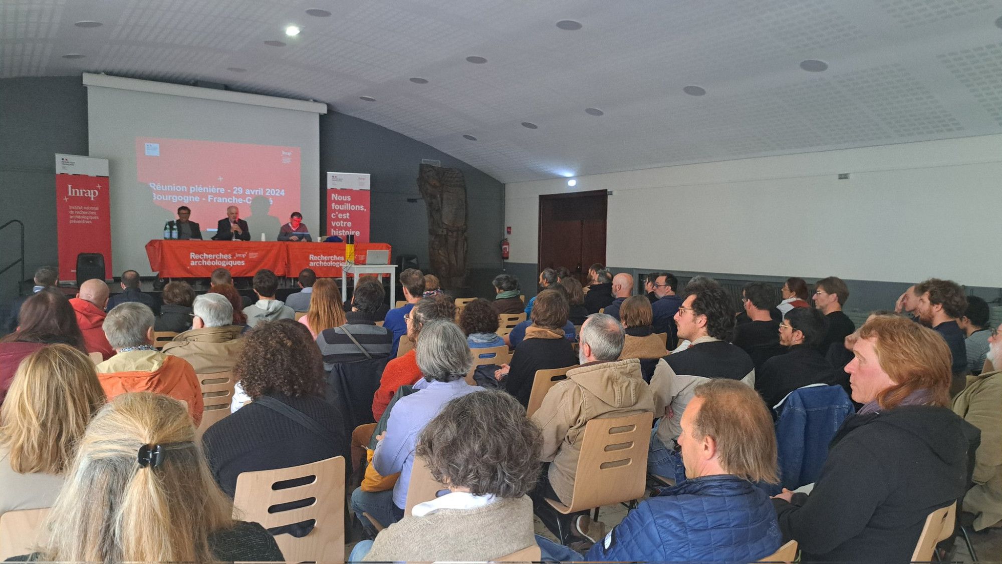 Photo d'une salle remplie de gens assis, avec en font trois hommes assis à une table sur une estradre