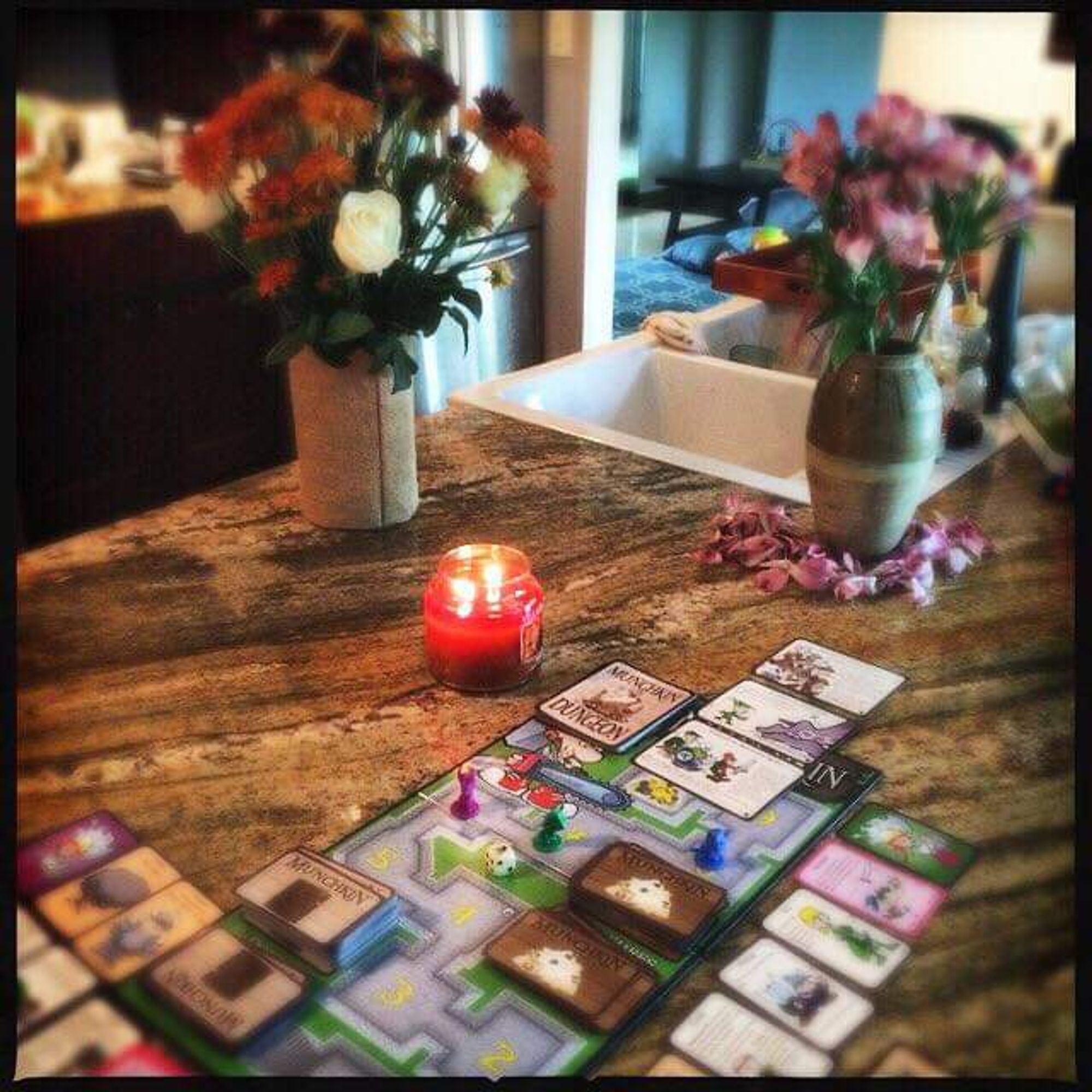 View of a countertop with two bouquets of flowers in vases, one with petals placed around it, a lit candle, and cards from a game of Munchkin.