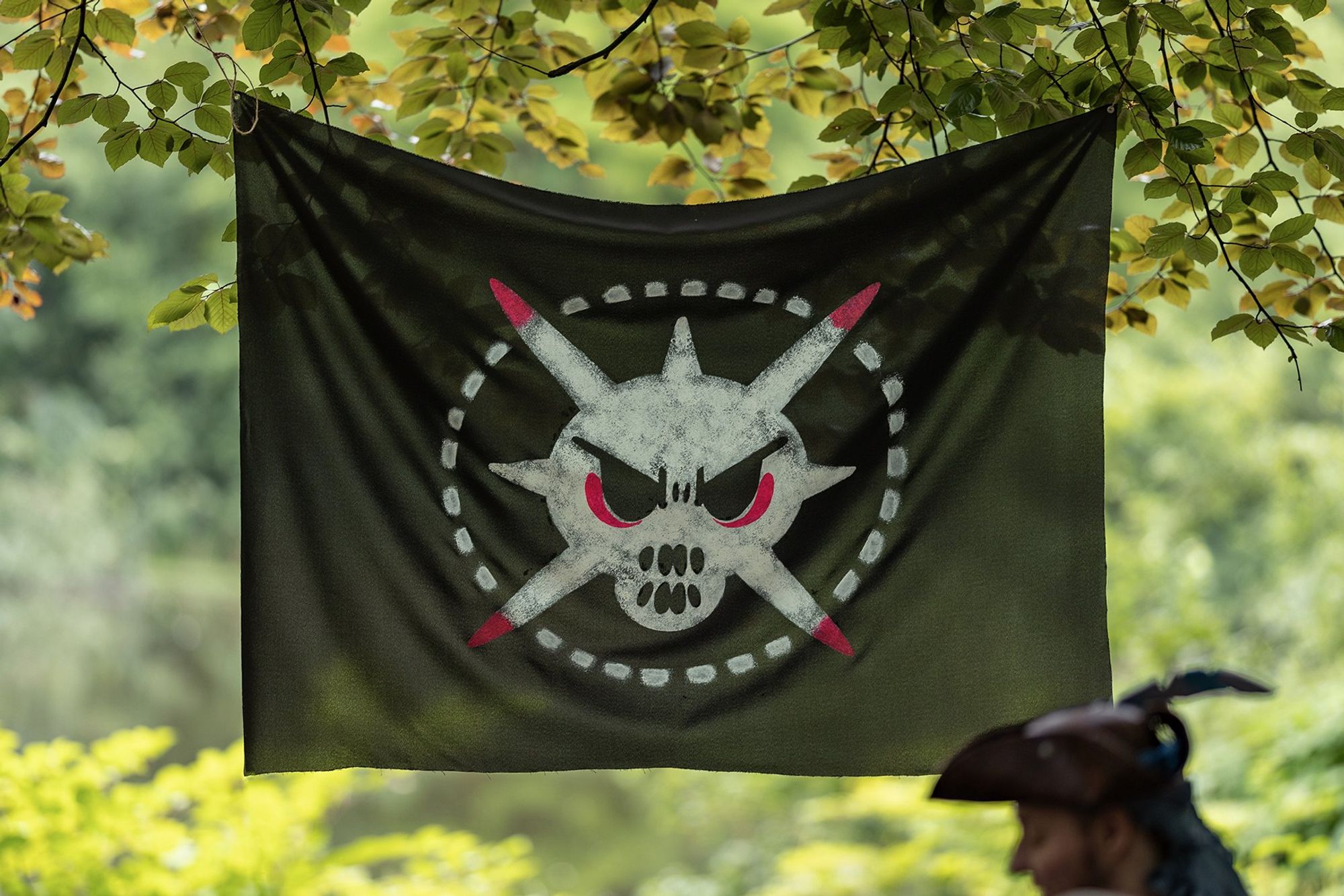 Eine schwarze Piratenflagge mit weißem Druck hängt in den Ästen eines Baumes. Das Motiv ist an das Pokémon Myridaor angelehnt. Unscharf davor steht jemand mit Piratenhut