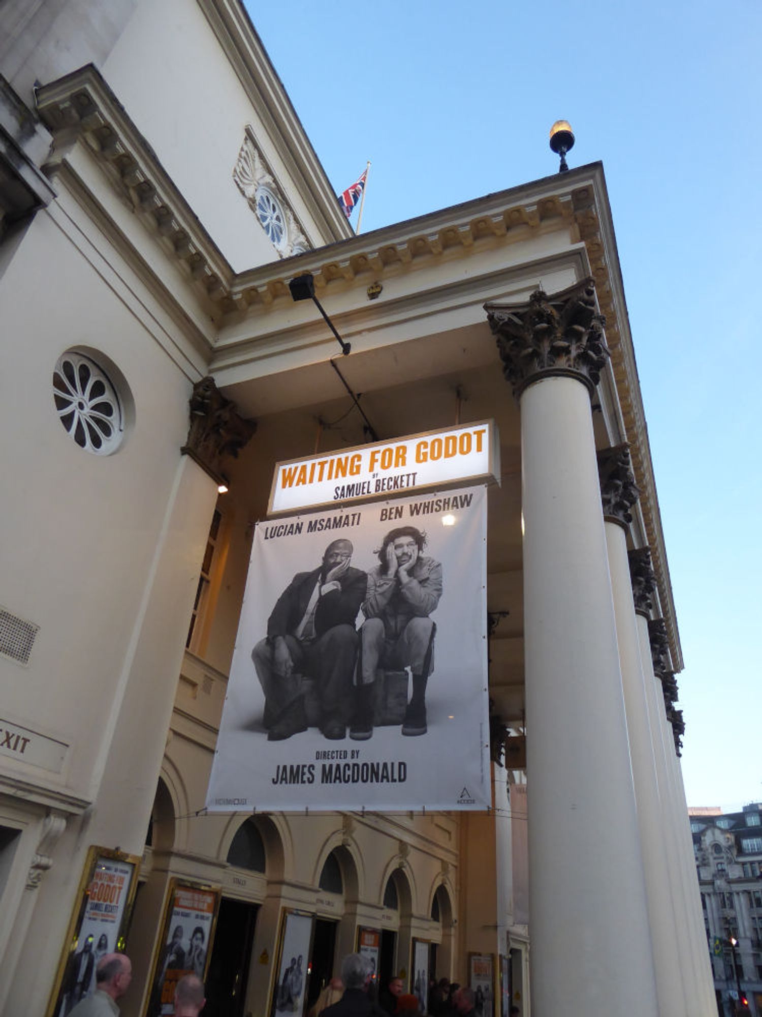 Poster hanging on side of white, pillared portico of theatre, showing black and white photo of Lucian Msamati and Ben Whishaw, seated. Early evening.