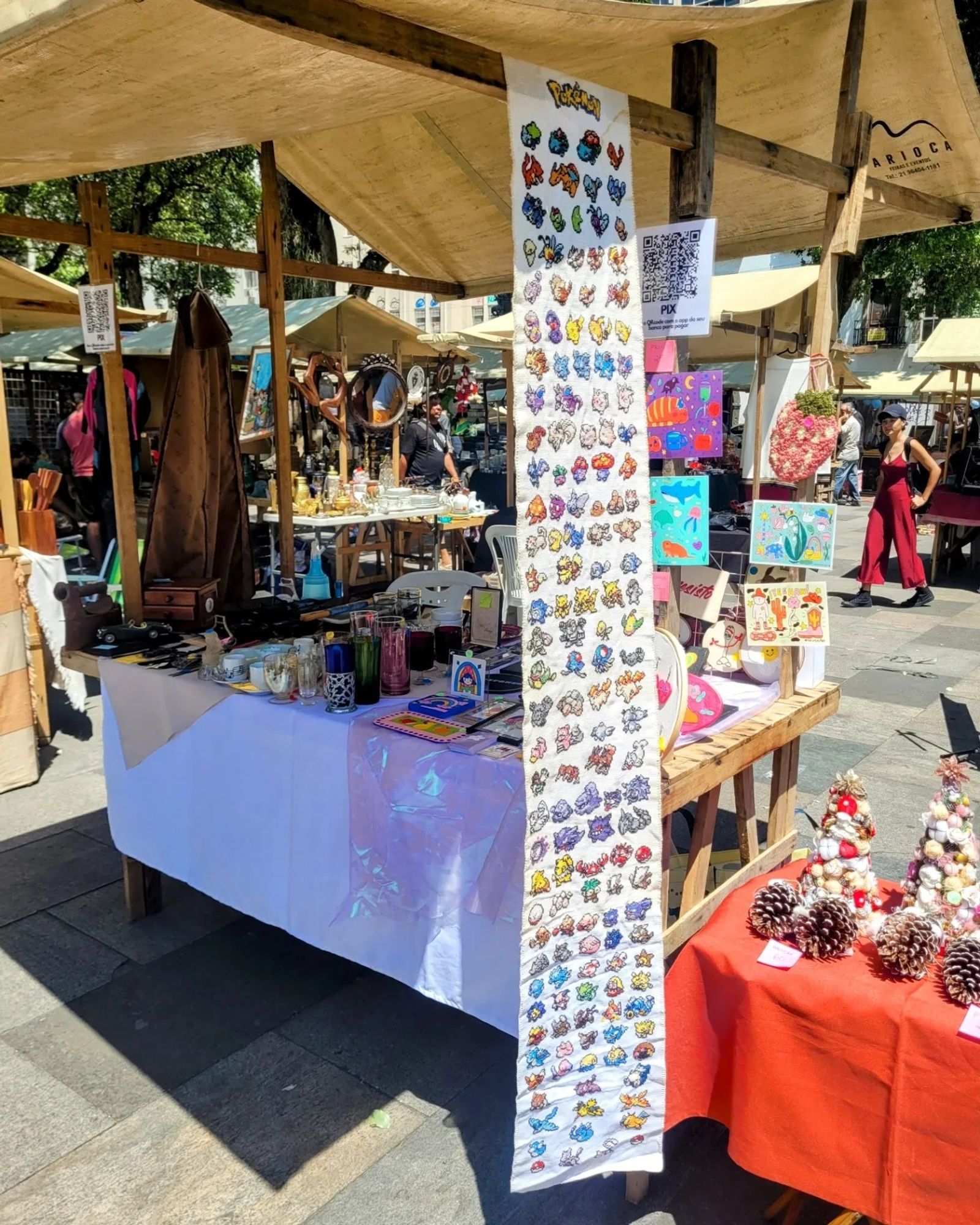 foto a distância da mesa da feira. ao lado, uma mesa mais baixa com decorações e artesanatos de natal feitos pela minha mãe. tem mini árvores decoradas e mini pinhos de decoração. o bordado gigante da primeira geração de pokemons feito pela julia está pendurado na barraca. ao fundo dá pra ver algumas das minhas ilustrações impressas e punch needle da julia. ainda mais ao fundo, antiguidades diversas vendidas pelo pai dela, flavio.