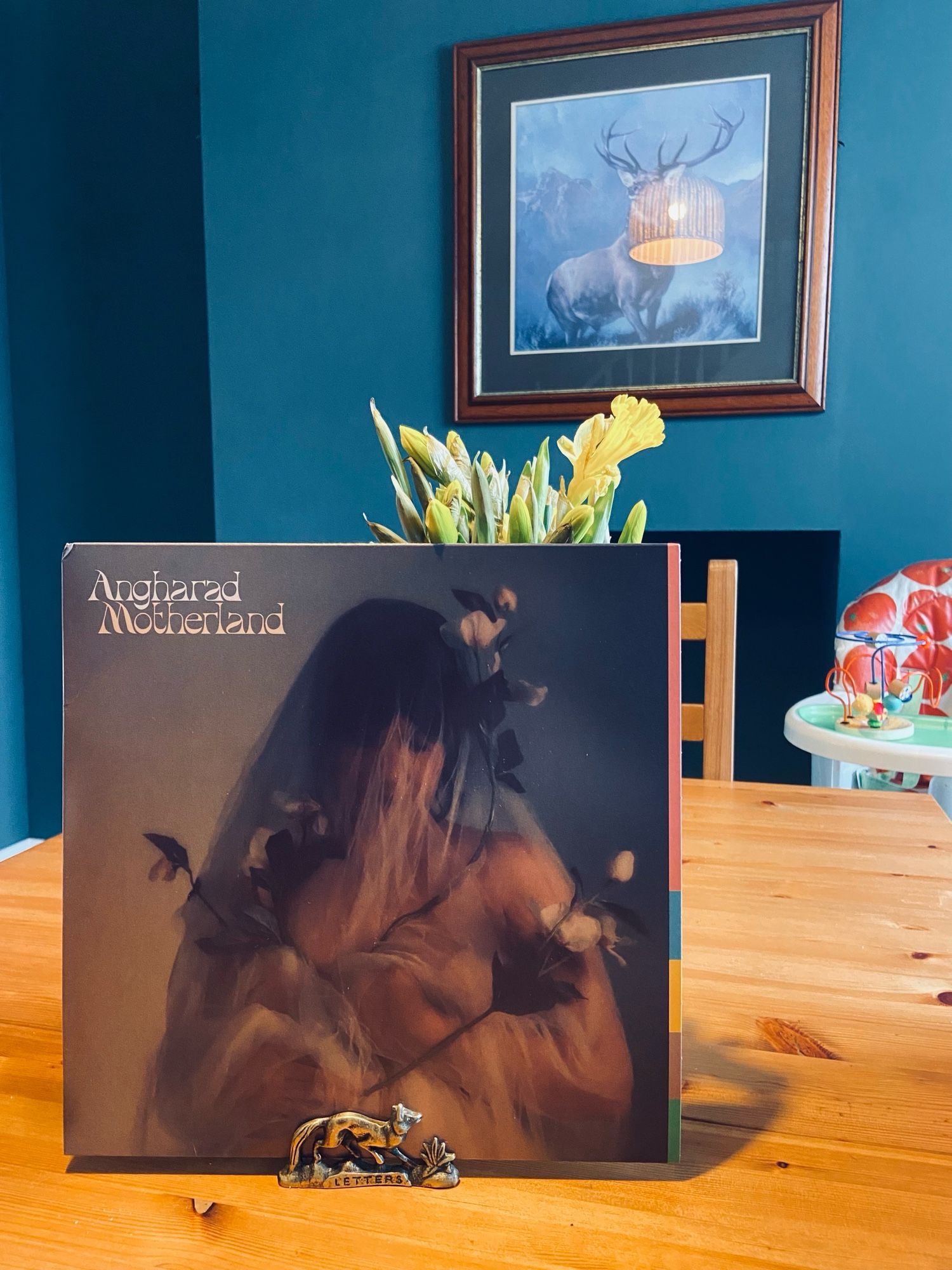 A vinyl record propped up against a vase of daffodils on a table in a blue room. The cover says ‘Angharad Motherland’ and has a photograph of Angharad beneath the writing.