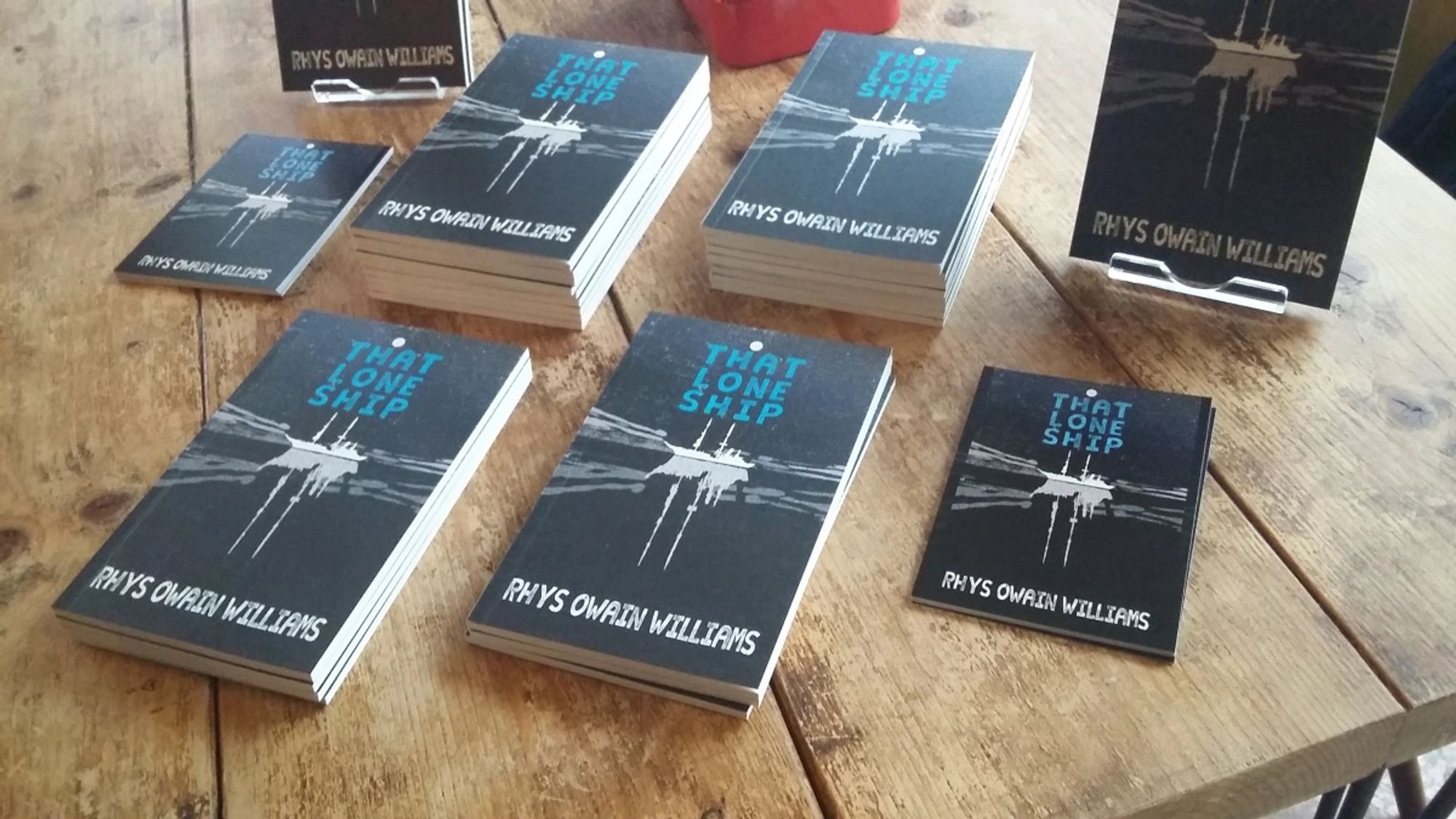 Stacks of That Lone Ship at the launch event. The cover has a white silhouetted ship on a black sea with the title of the book above it and my name below it.