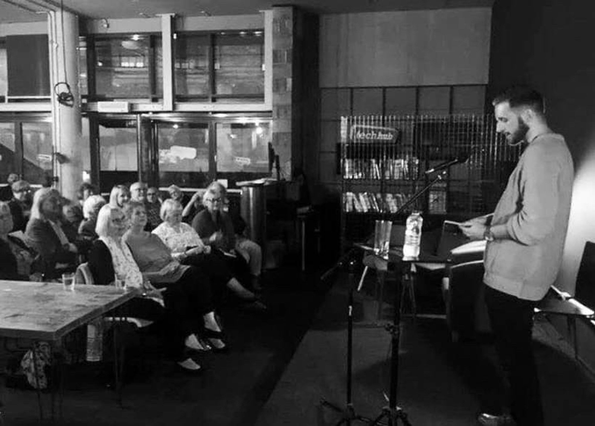 Me reading from That Lone Ship to a crowd at TechHub Cafe in Swansea in September 2018. I am on a stage with a microphone and still have hair on my head.