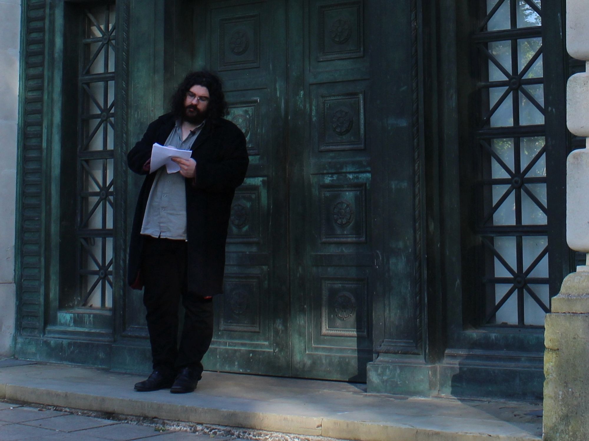 Christopher Cornwell stands in front of the ornate doors of Swansea’s Guildhall. He has long dark hair and a beard and is writing on a piece of paper.