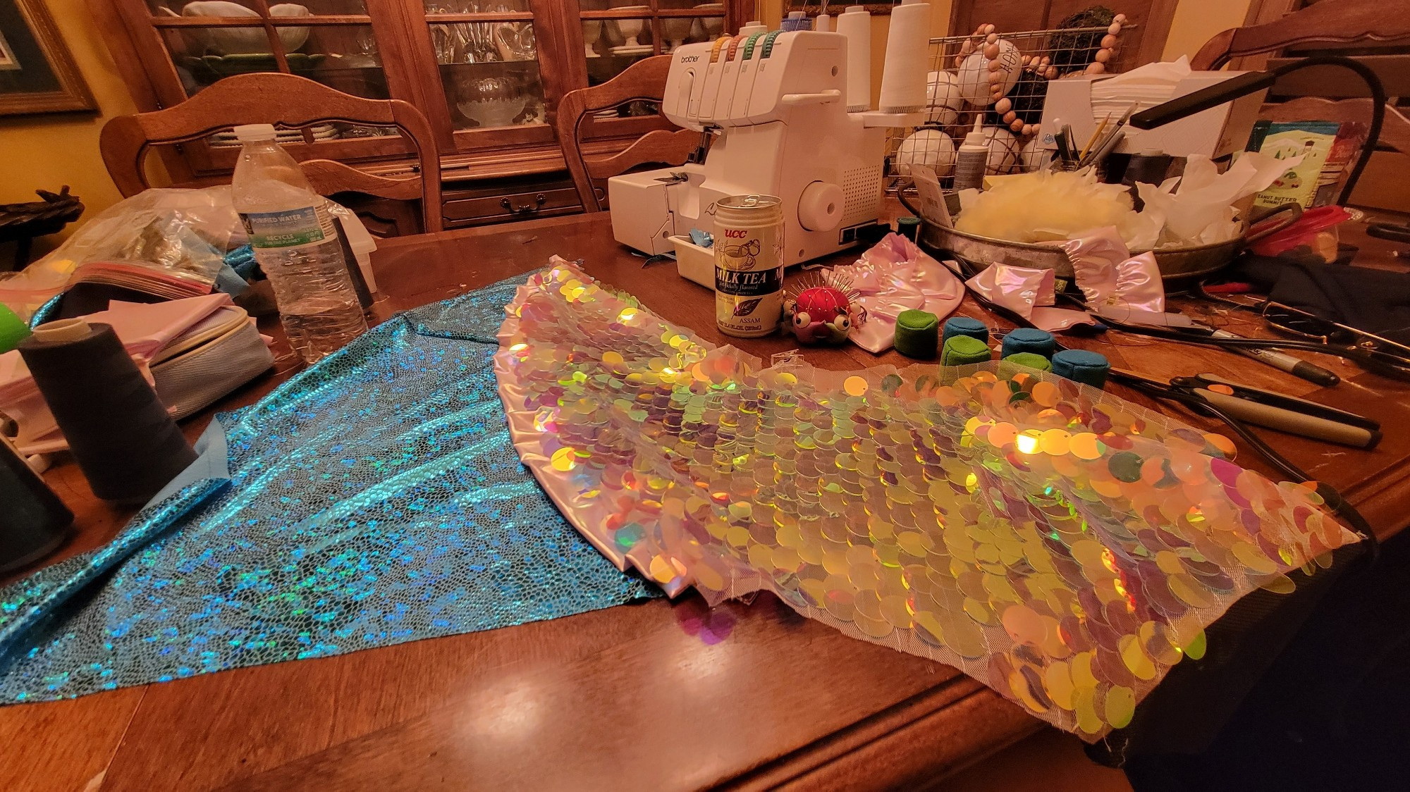 Wip shot of leg of coatume featuring 1" sequin fabric, pink holo ruffles, and blue/teal scaled foil spandex. Serger and other sewing accessing around