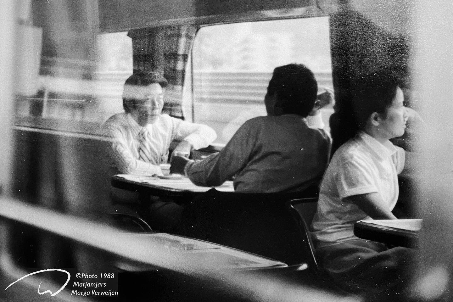 Black and white image of people sitting at tables in a high speed Shinkansen train in Japan taken through a window with reflections, 1988.