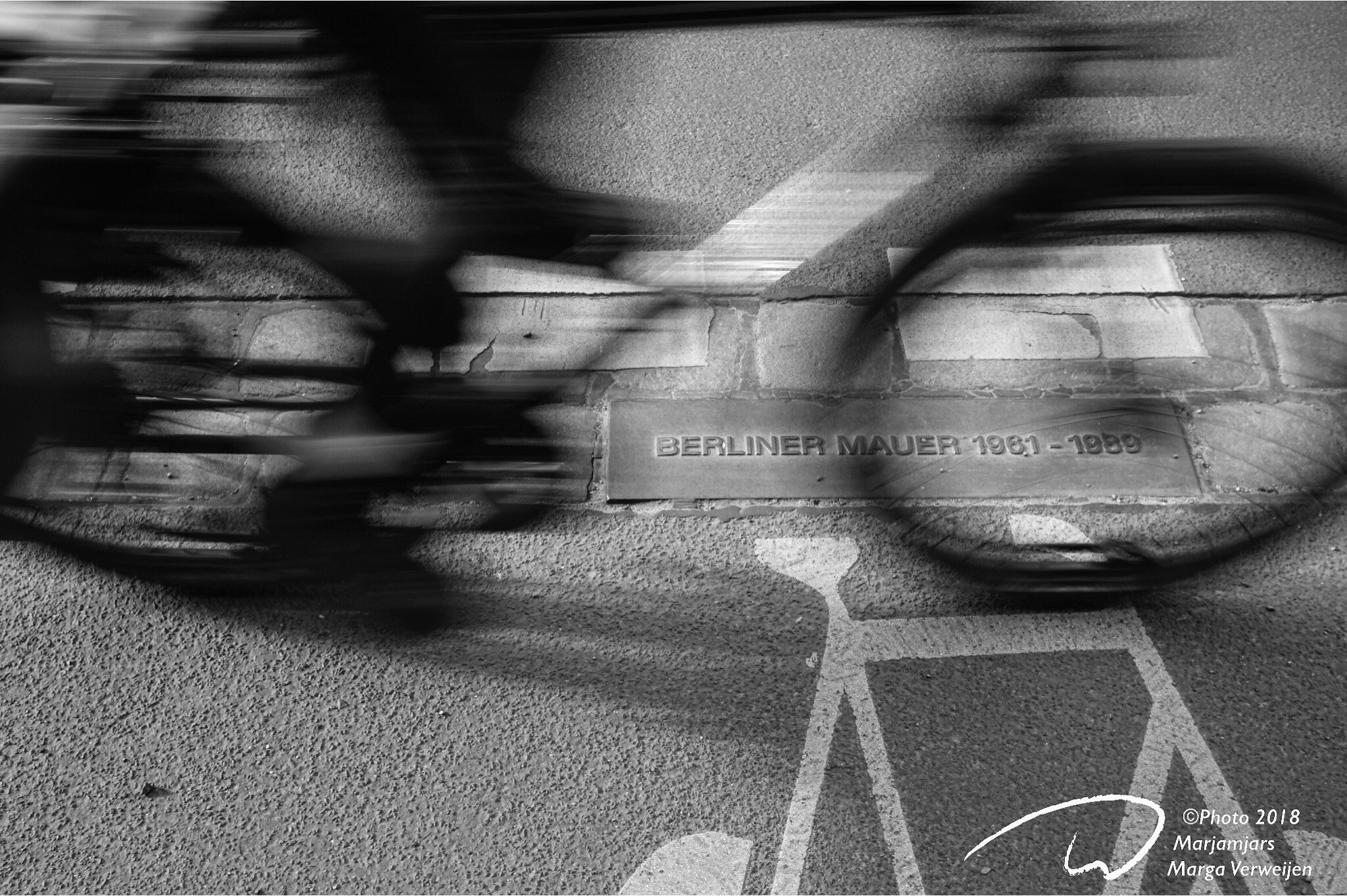 A moving bicycle on a the streets of Berlin. On the ground a double row of cobble stones mark the former route of the wall in Berlin. Also shows a metal plate ‘Berliner Mauer 1961 a 1989’