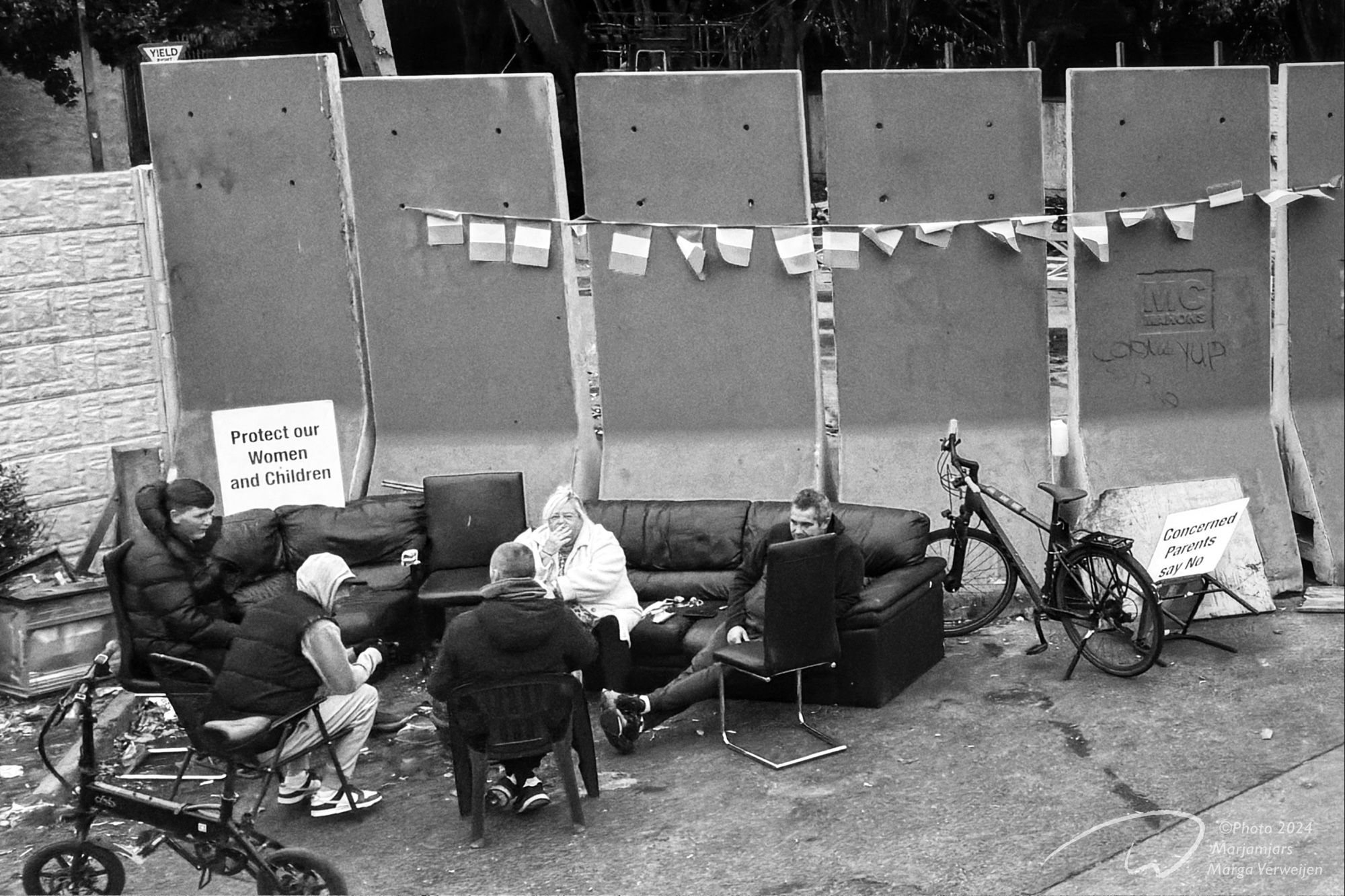 Months after the riots, protesters at proposed asylum centre location in Coolock are in silent protest. Some old furniture bicycles and people in front of the barricaded location. Irish flags hang on the wall Signs that read ‘Protect our women and children’ and ‘Concerned parents say no’ on display.