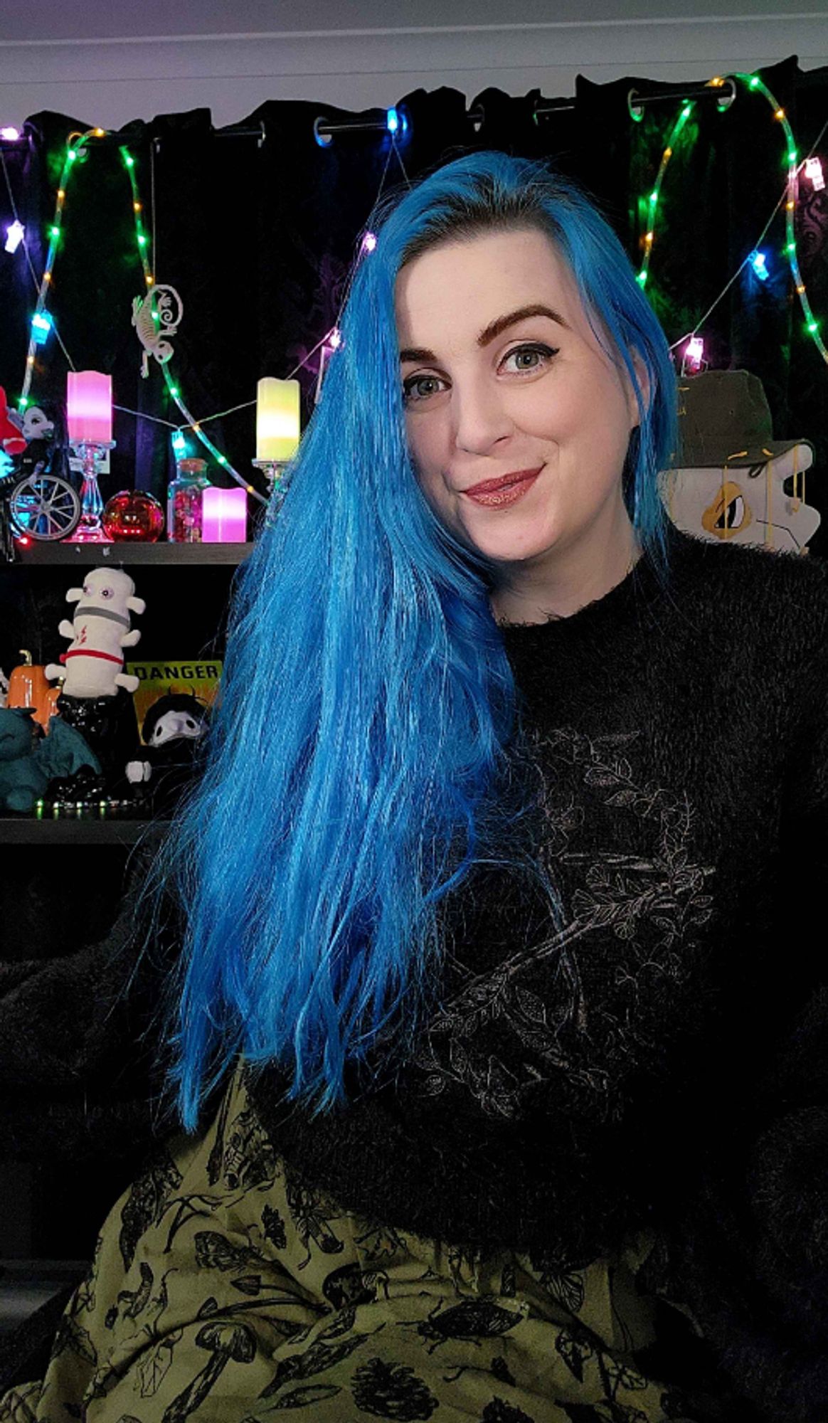 A photograph of Dragancaor, a white nonbinary femme streamer with green eyes & long blue hair. They are wearing an olive green dress & a black fluffy jumper (pullover), both with hedgewitch designs, by Killstar