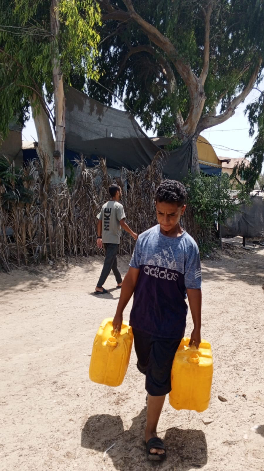 Mahmoud carries 2 jugs of water. The exertion is lined in his face