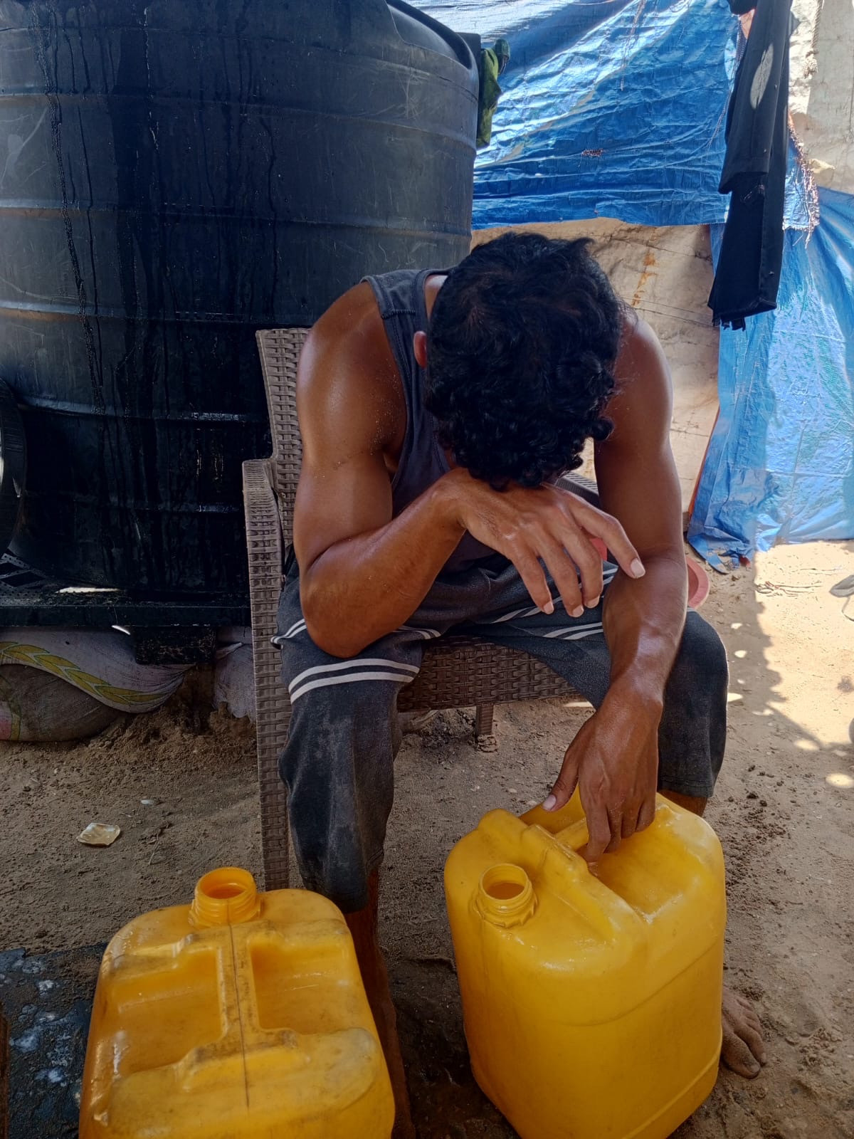 Abdullah sits slumped in a chair, resting his head on the back of his hand. He is covered in sweat & visibly exhausted. Before his are 2 large yellow jugs for water 