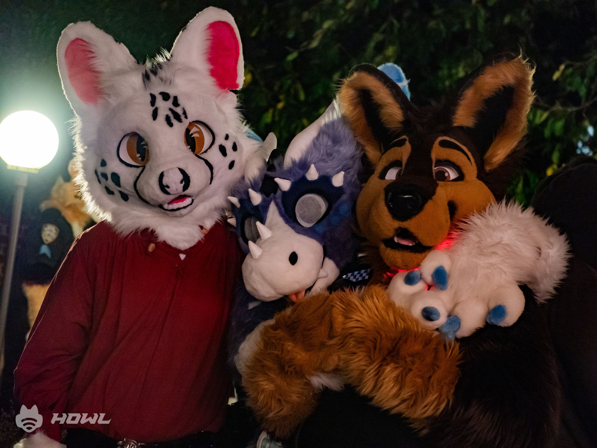 Three fursuiters hugging each other: A serval, a dutch angel dragon and a wolf.