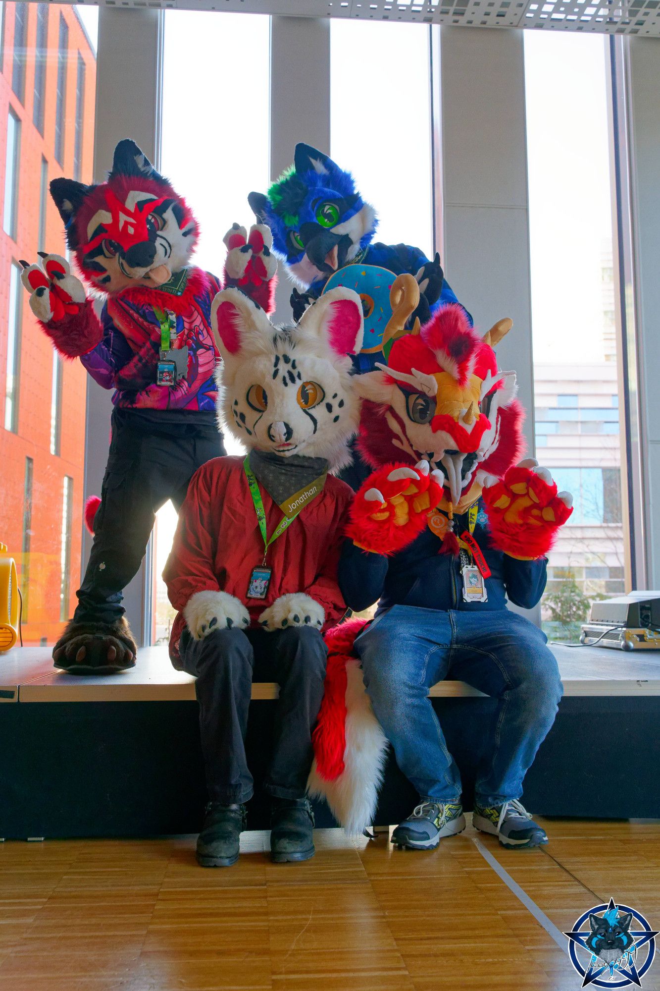 A group photo showing four fursuiters: A red and white dog, a blue and white bird, a white serval and a red and white dragon.