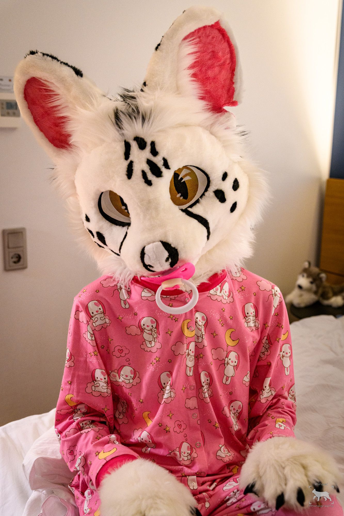 Photo of a white serval partial fursuiter wearing pink pyjamas with a pink pacifier in their mouth.