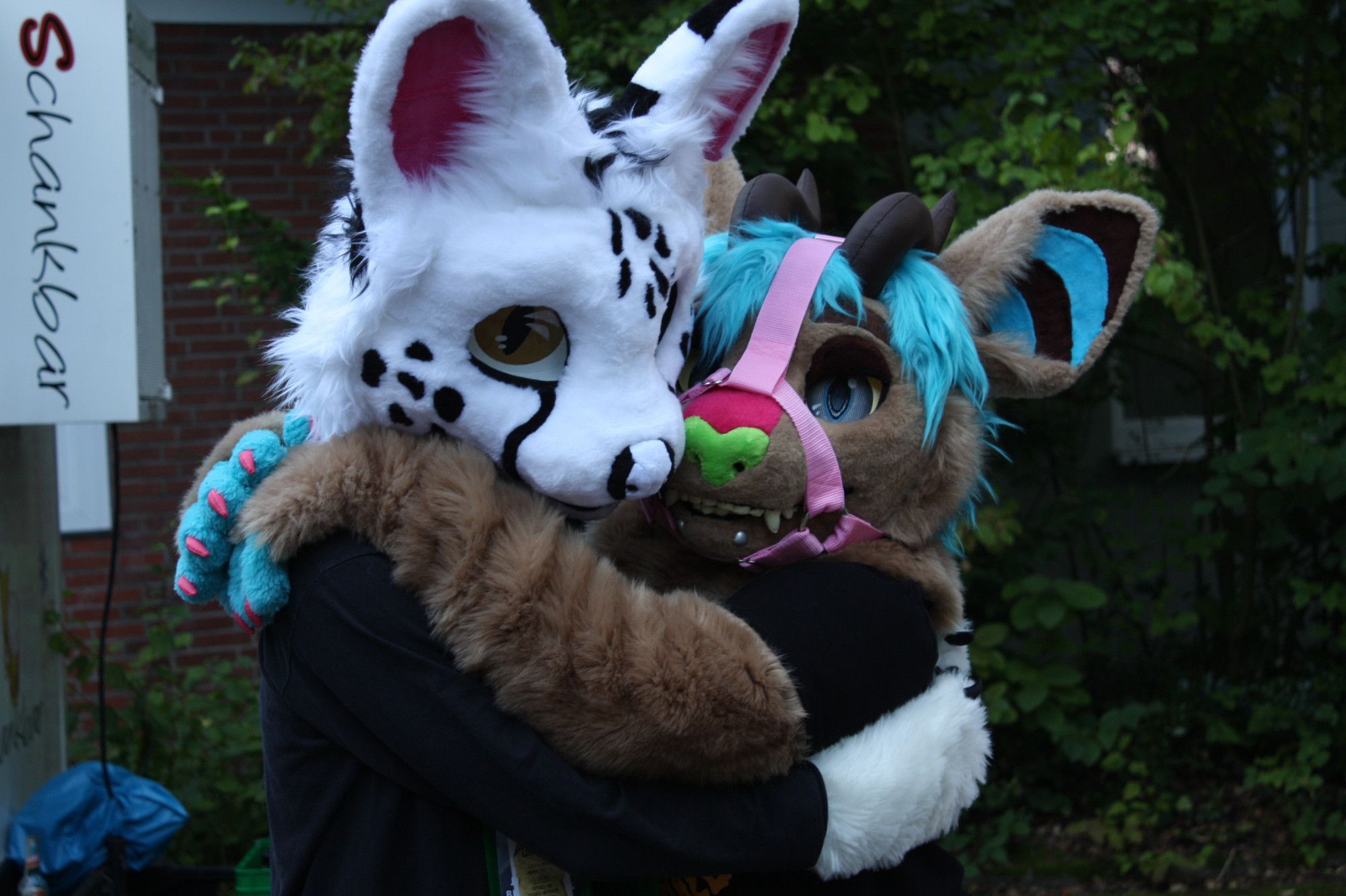 Photo of a white serval fursuiter and a cat/bunny/demon hybrid fursuiter hugging each other.