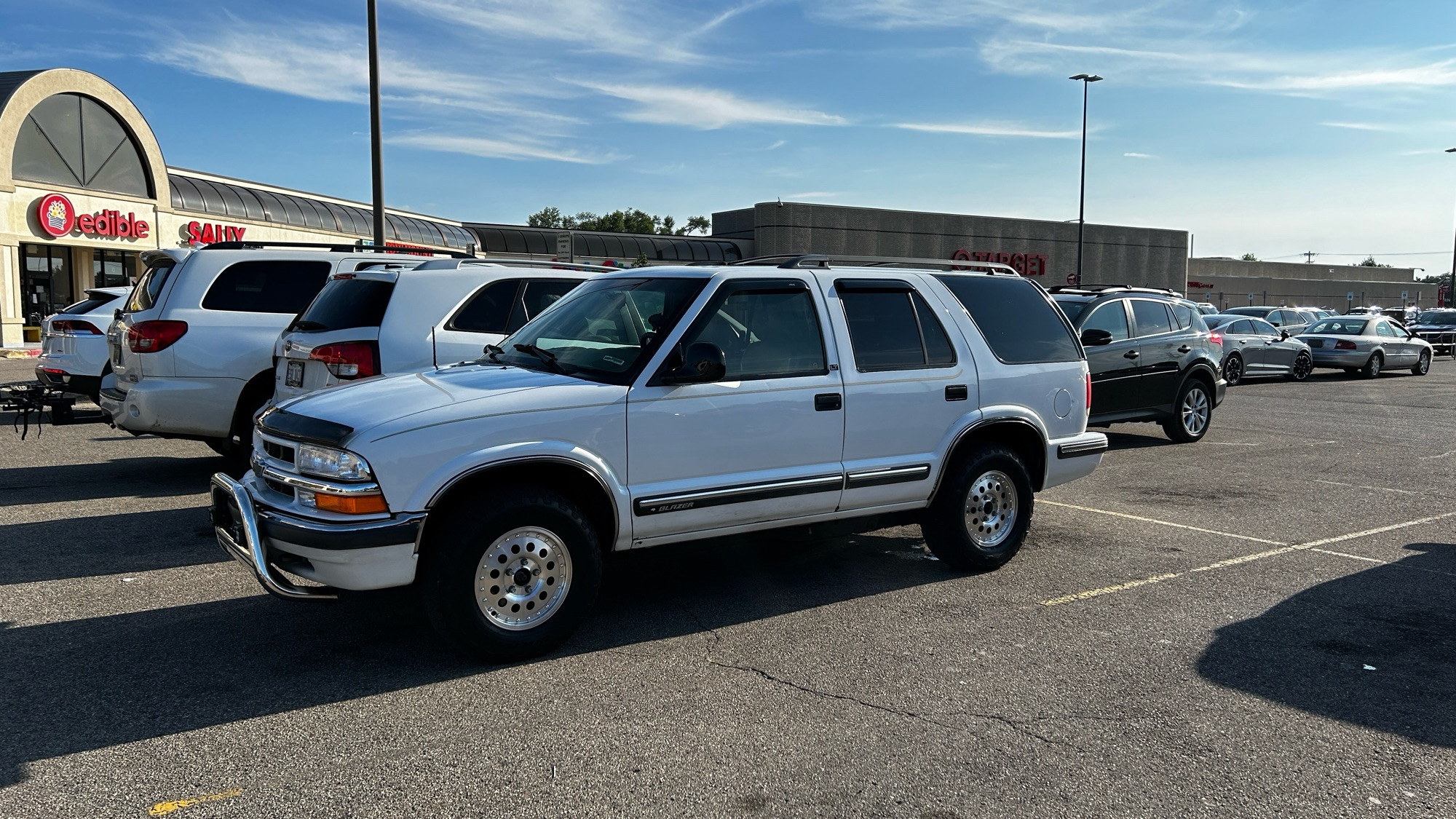 A very clean, shiny even, white 2000s-era Blazer (2nd gen of the s-10 blazer line according to Wikipedia because wow confusing), with shiny steel wheels