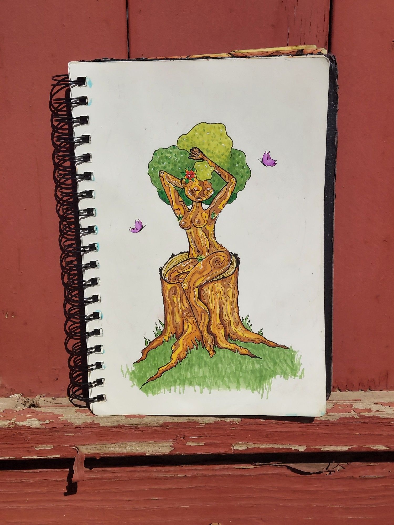 A color drawing of a naked tree lady. She's got skin that looks like bark with different shades of brown,orange and yellow a leafy green afro. she is siting on a tree stump pin up style with purple butterfly's around her