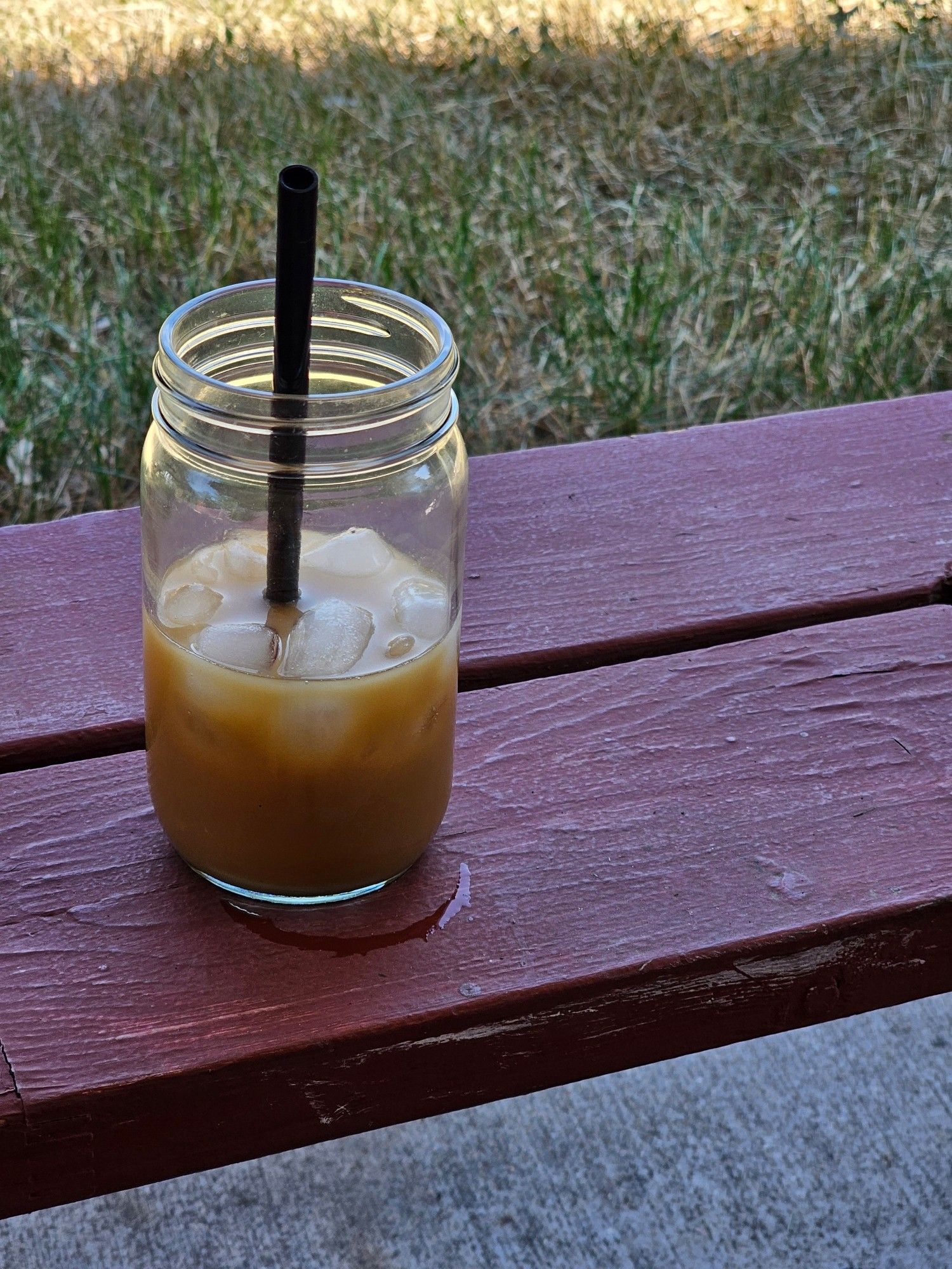 Adolinntook this pocture while enjoying their moring coffee. Pictured is an iced Oat milk dirty chai, in a jar sitting on a red bench. There's grass in the back ground.