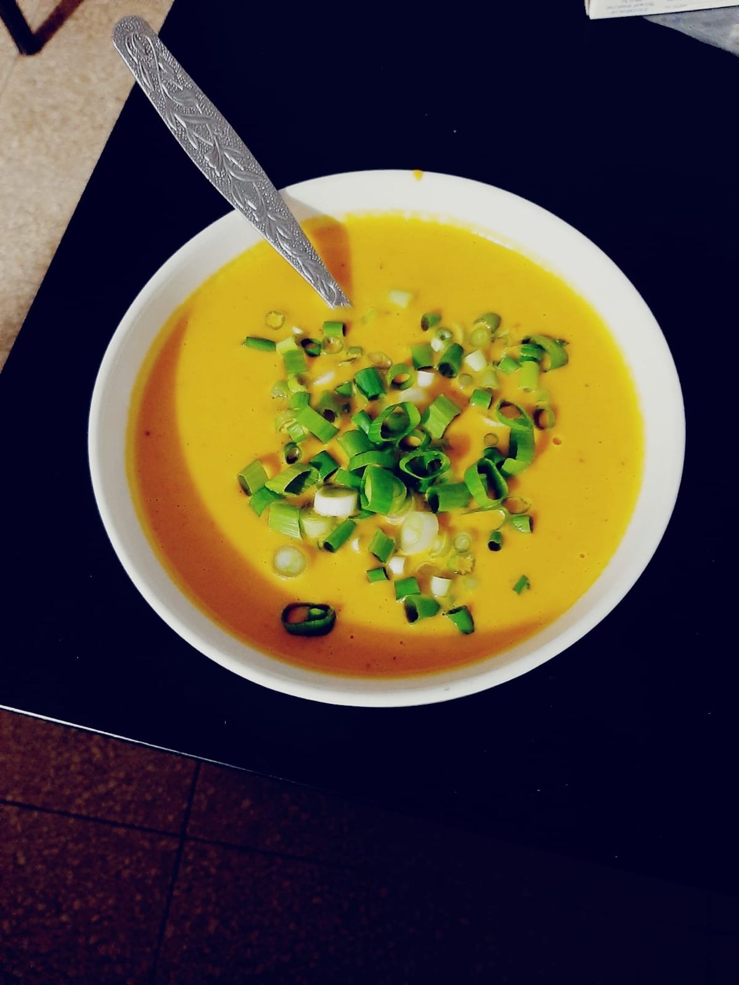 A picture of a pumpkin cream soup, the only thing I'm capable of making in the kitchen. It is almost neon orange with a bit of green onion chopped on top, in the medium sized white bowl with the spoon dipping inside on the black coffee table.