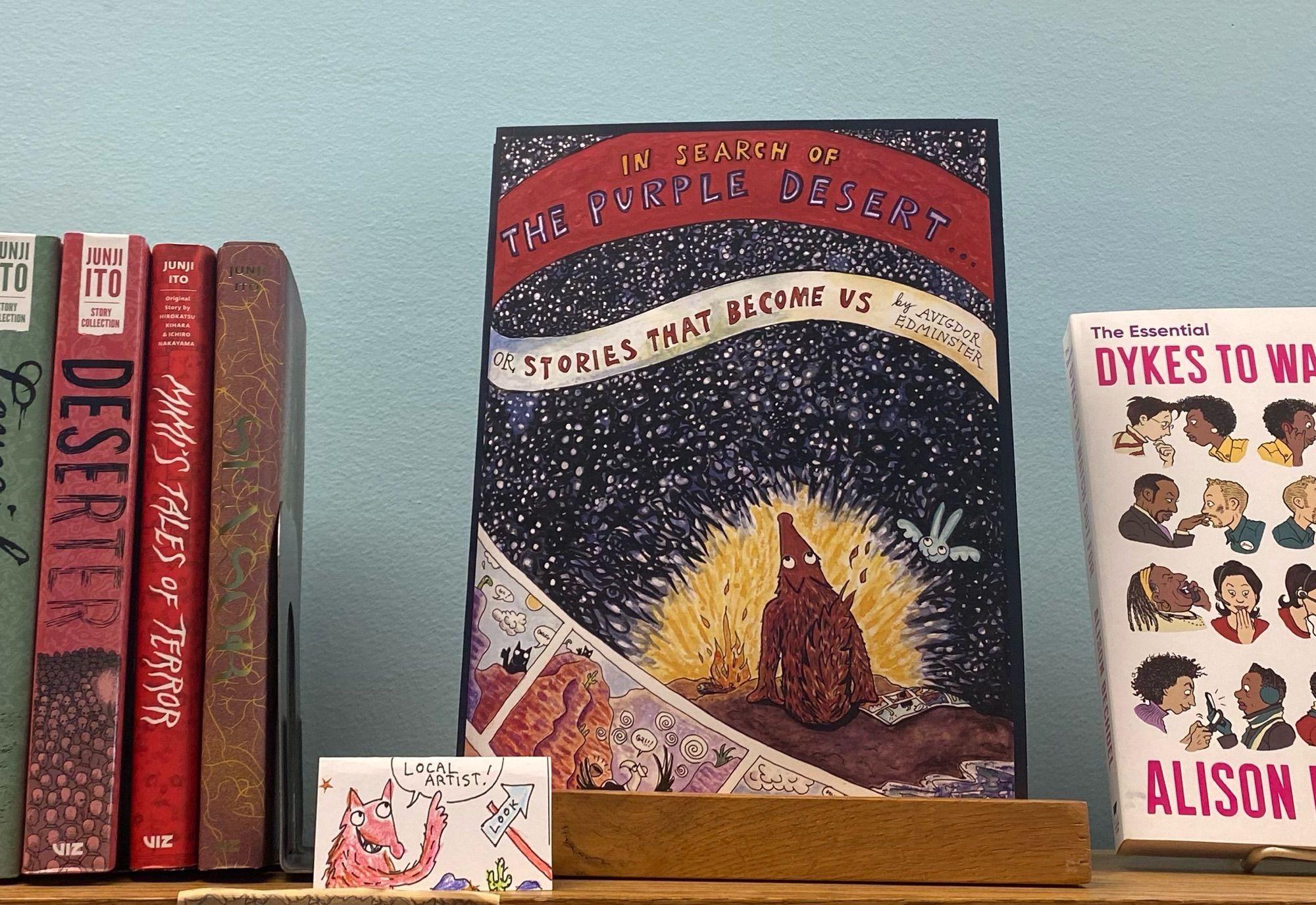 Book cover of graphic novel “In Search of the Purple Desert” - sitting on book shelf. Cover shows red coyote and floating blue bird looking up into the night sky, in front of blazing bonfire. Red Coyote gazes at the thousands of stars, representing stories of our ancestors. In lower left cover, a preview comic strip of inside of graphic novel with desert landscapes, with Eagle, snake, and aloe plants.