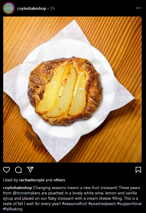 A screenshot image from @coylesbakeshop on Instagram. The image is of a round, pear croissant placed on a white dish on top of a wooden table surface. The fruit pastry is golden browned and caramelized. The post's caption reads, "Changing seasons means a new fruit croissant! These pears from @tonnemakers are poached in a lovely white wine, lemon and vanilla syrup and placed on our flaky croissant with a cream cheese filling. This is a taste of fall I wait for every year! #seasonalfruit #poachedpears #supportlocal #fallbaking"