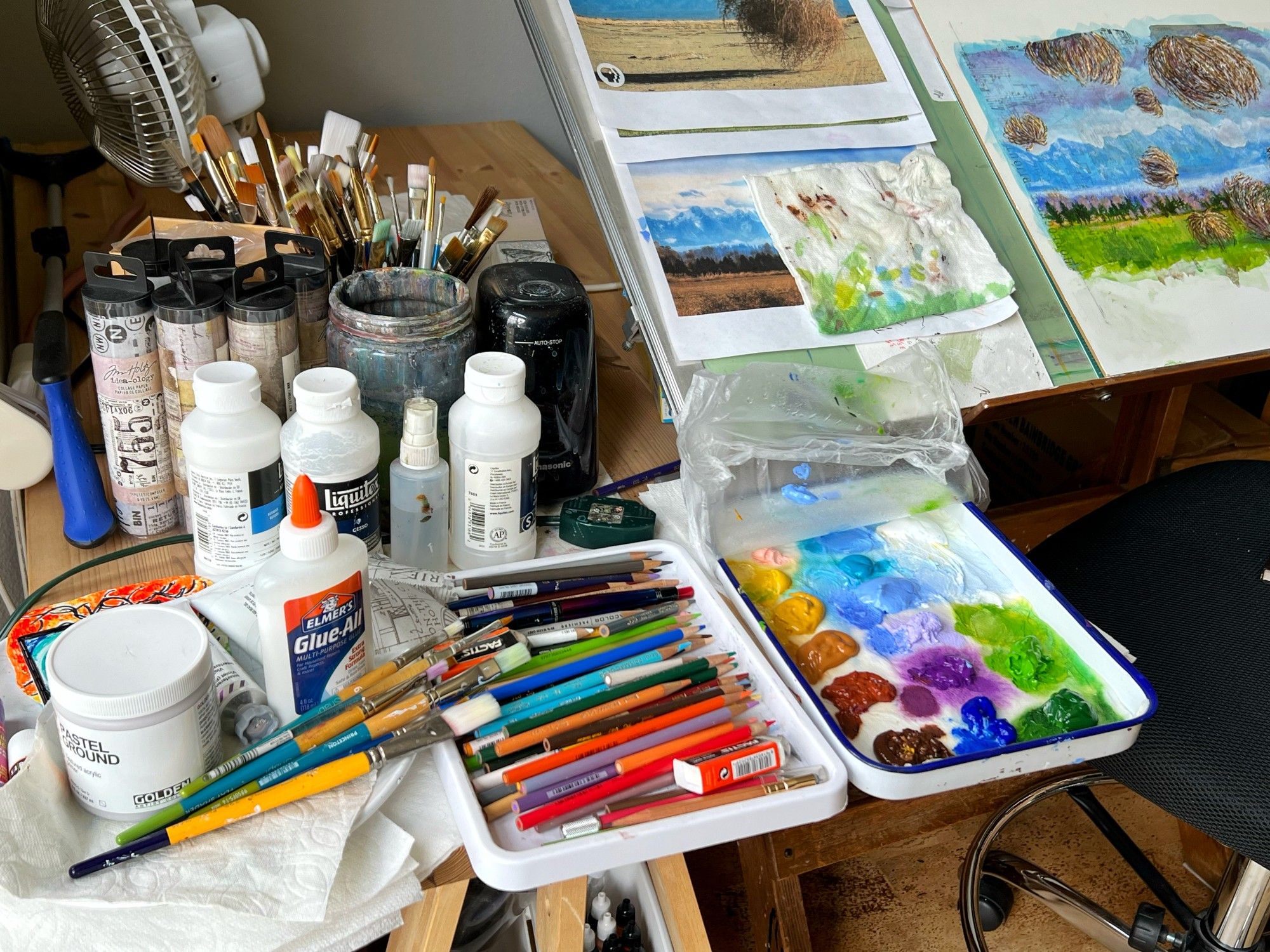 A very cluttered art cart sits on the left side of a drawing table with print-outs and paintings on it. A palette tray with wet paints sits next to a tray filled with colored pencils, and jars of brushes sit behind it all.