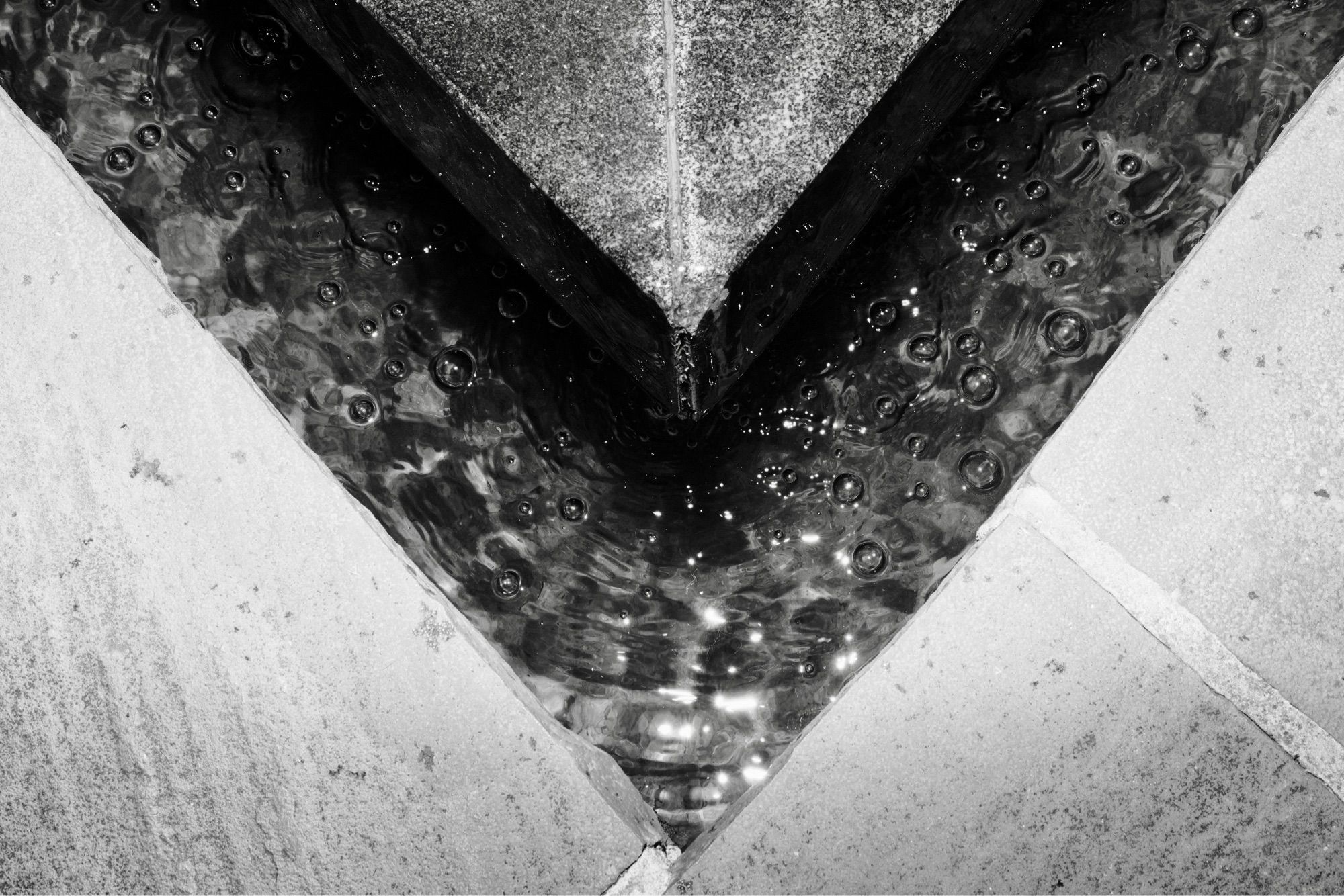 A v-shaped channel separates two halves of a fountain. Water droplets sparkle in mid-collapse on the surface of a flow through the channel.