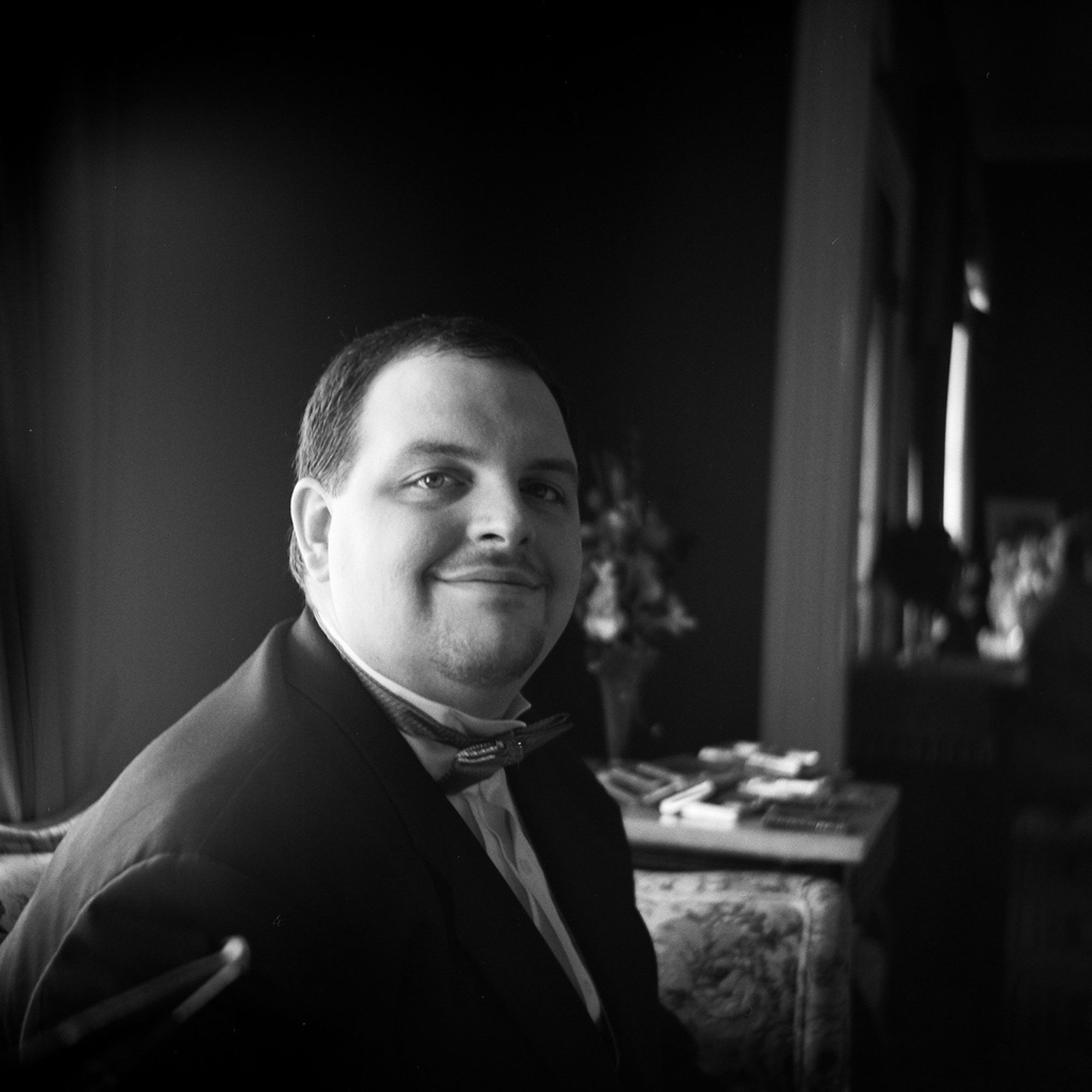 A groom in a tuxedo sits in gentle light looking at the camera. His expression is a small, peaceful smile.