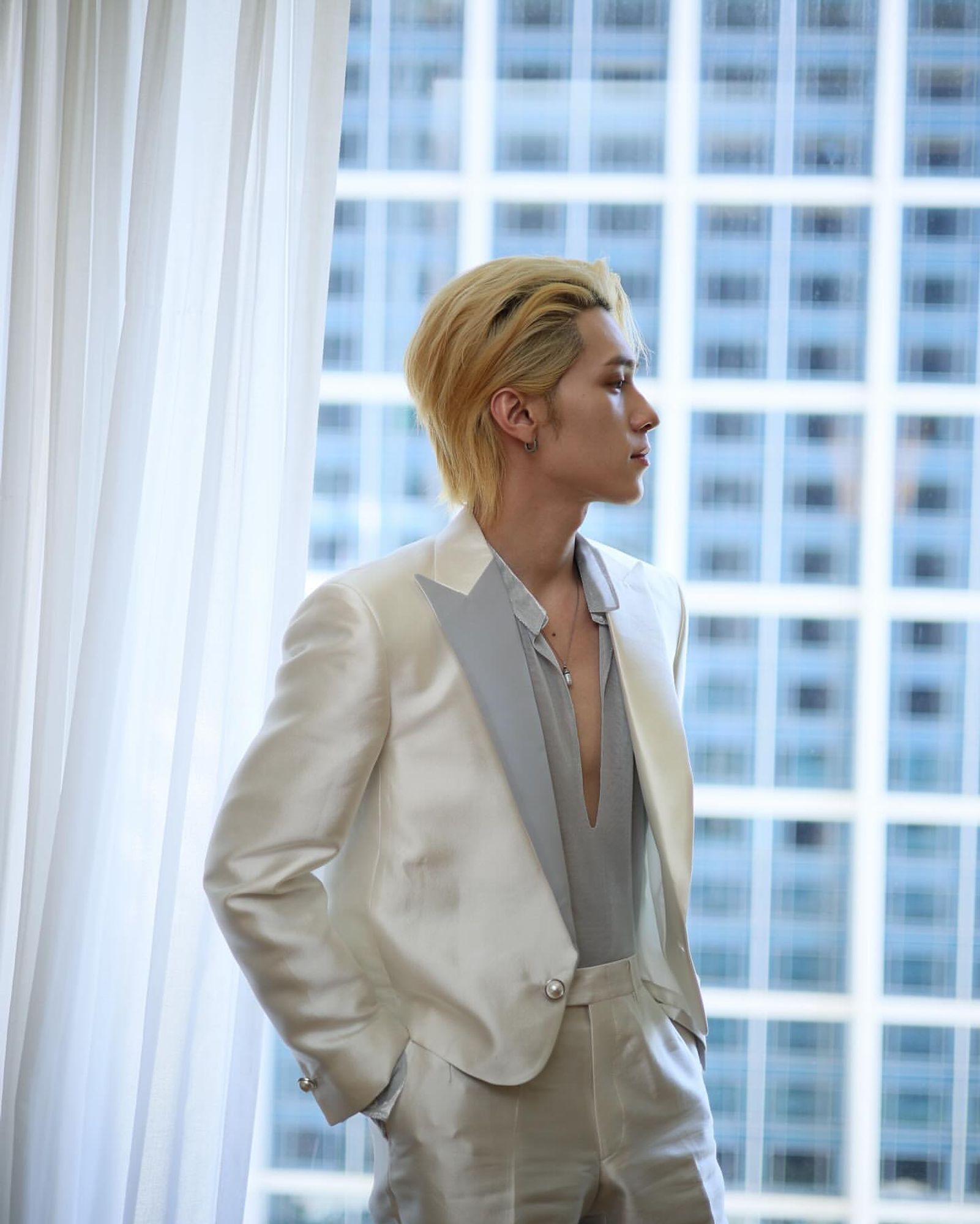 side profile photo of hendery wearing a prince-like white and light blue-ish grey suit with a plunging neckline and blonde hair