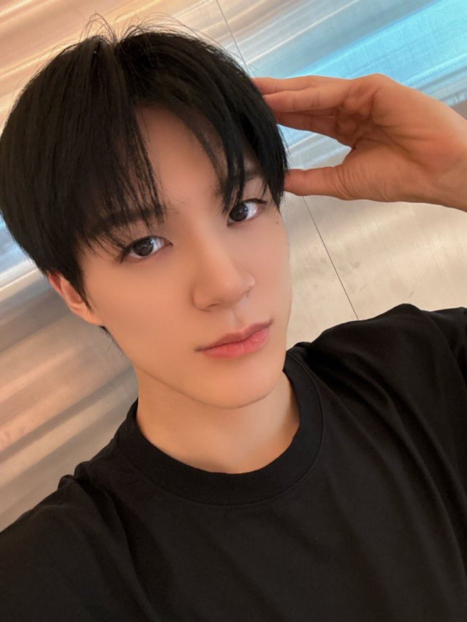 cute selca of a straightfaced jeno in a black shirt looking straight at the camera with a hand up doing the floppy ear puppy pose