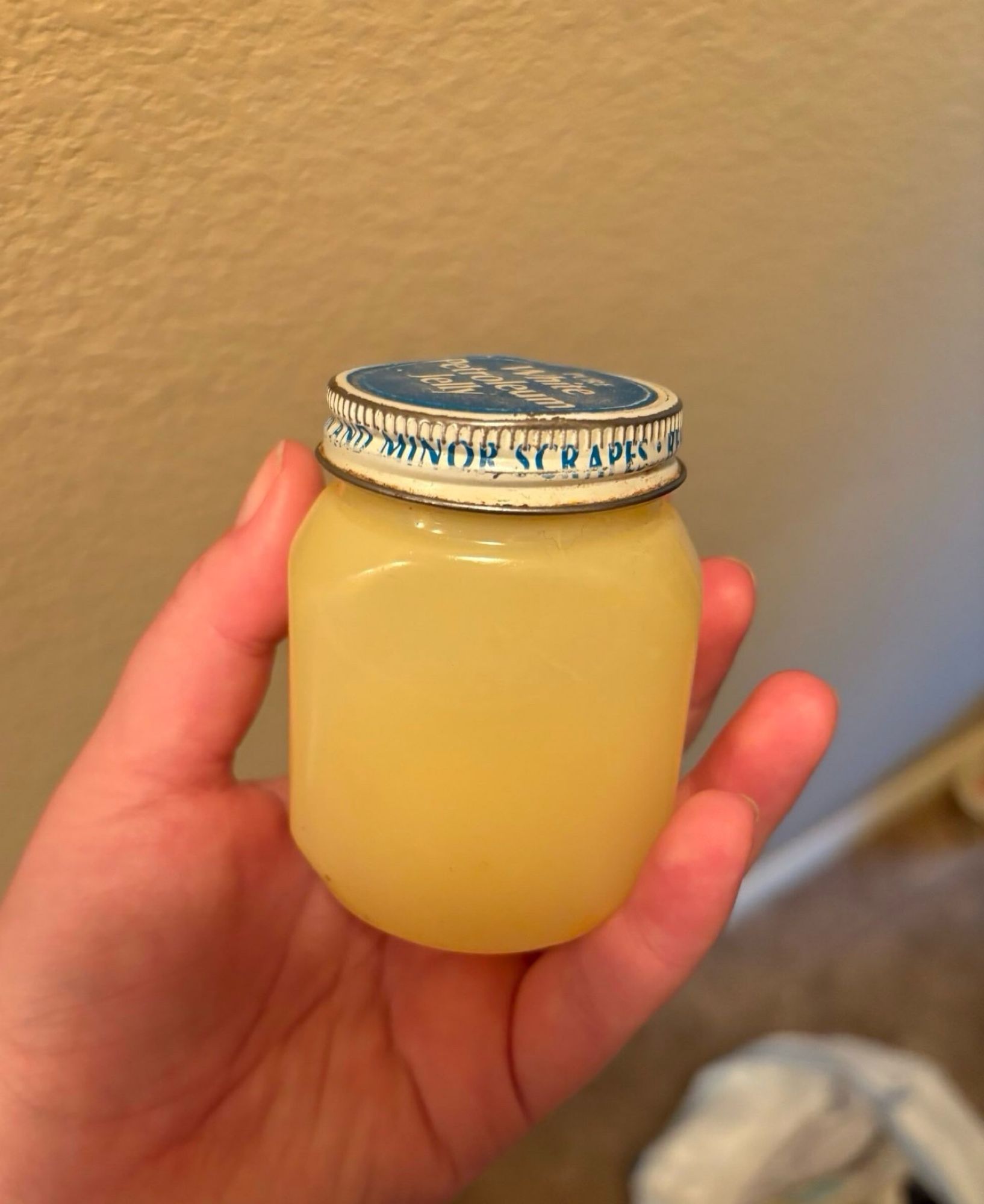 A left hand holding an old jar of petroleum jelly that says minor scrapes on the side of the lid with a very yellow discolored container