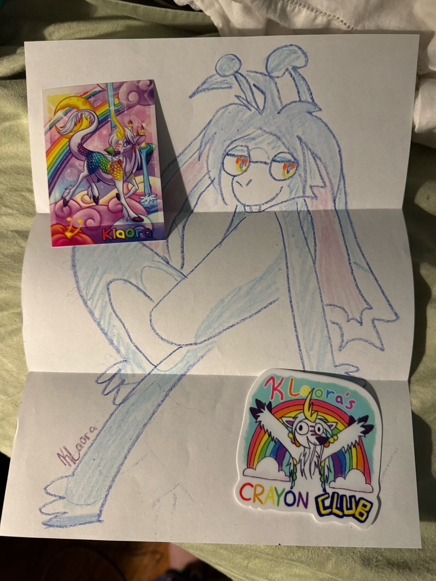 A crayon drawing of a blue alien bunny. Beside it is a sticker of a kirin in front of a rainbow, and Klaora’s very colorful artist business card.