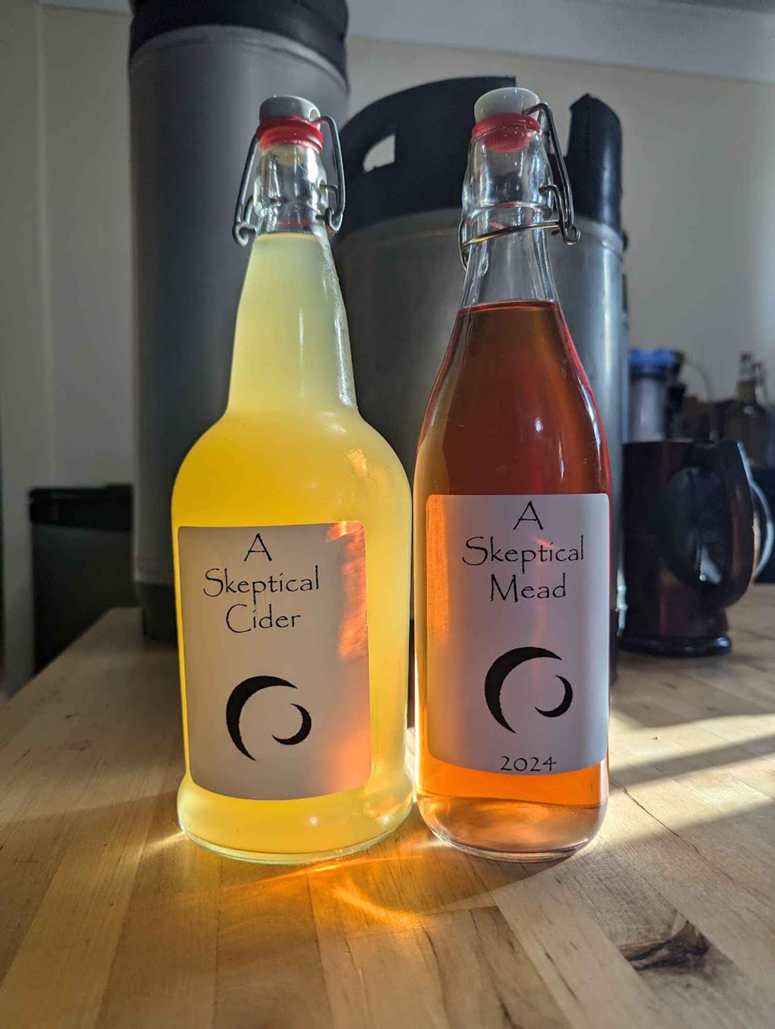 Two glass bottles on a wooden table, larger bottle reads "A Skeptical Cider" the smaller bottle reads "A Skeptical Mead". Two kegs can be seen in the background.