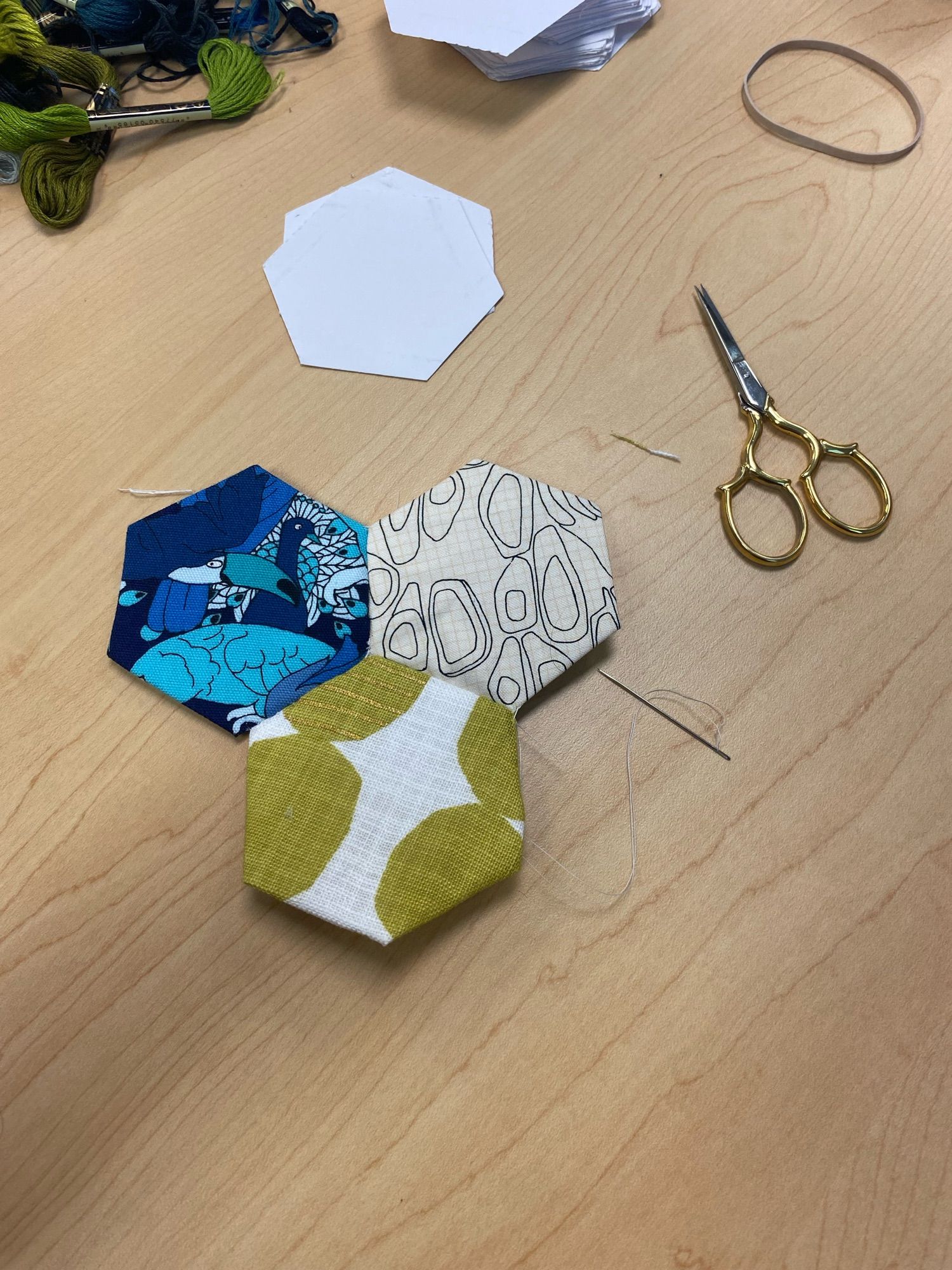 Three hex shaped quilt pieces in different colored fabrics sitting on a table next to a small pair of thread snippers and a threaded needle.