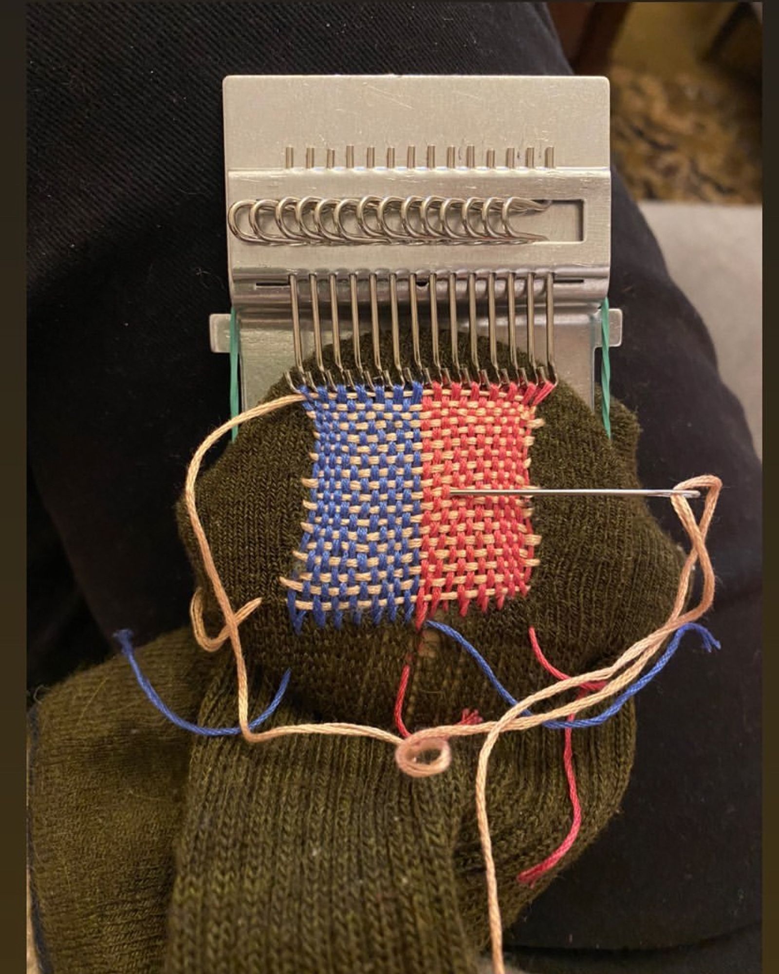 A metal darning loom attached to a green sock, with a patch in progress. Red, blue, and yellow thread woven to cover a hole in the heel of the sock.