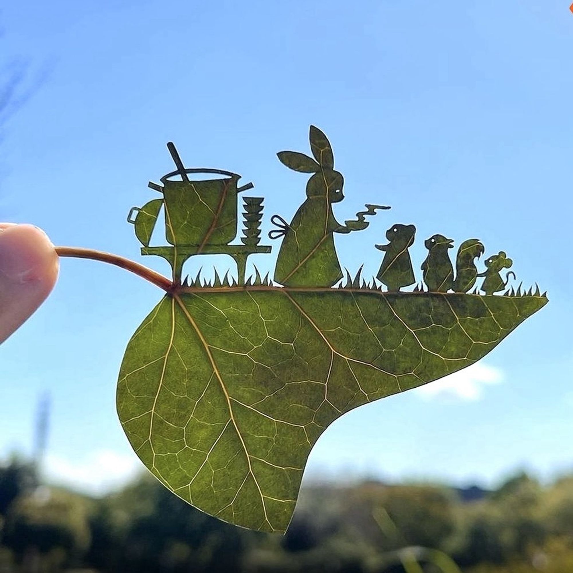 An ivy leaf is held horizontally in front of a blue sky.  The top half is carved, the bottom half is untouched.
The scene depicts a large pot on a low table with the lid leaning against the side of the pot and a stack of bowls also on the table.
Next to it stand a rabbit and row of smaller animals: a cat(?), a squirrel and a mouse.  The rabbit is handing out a steaming bowl to the first little one.