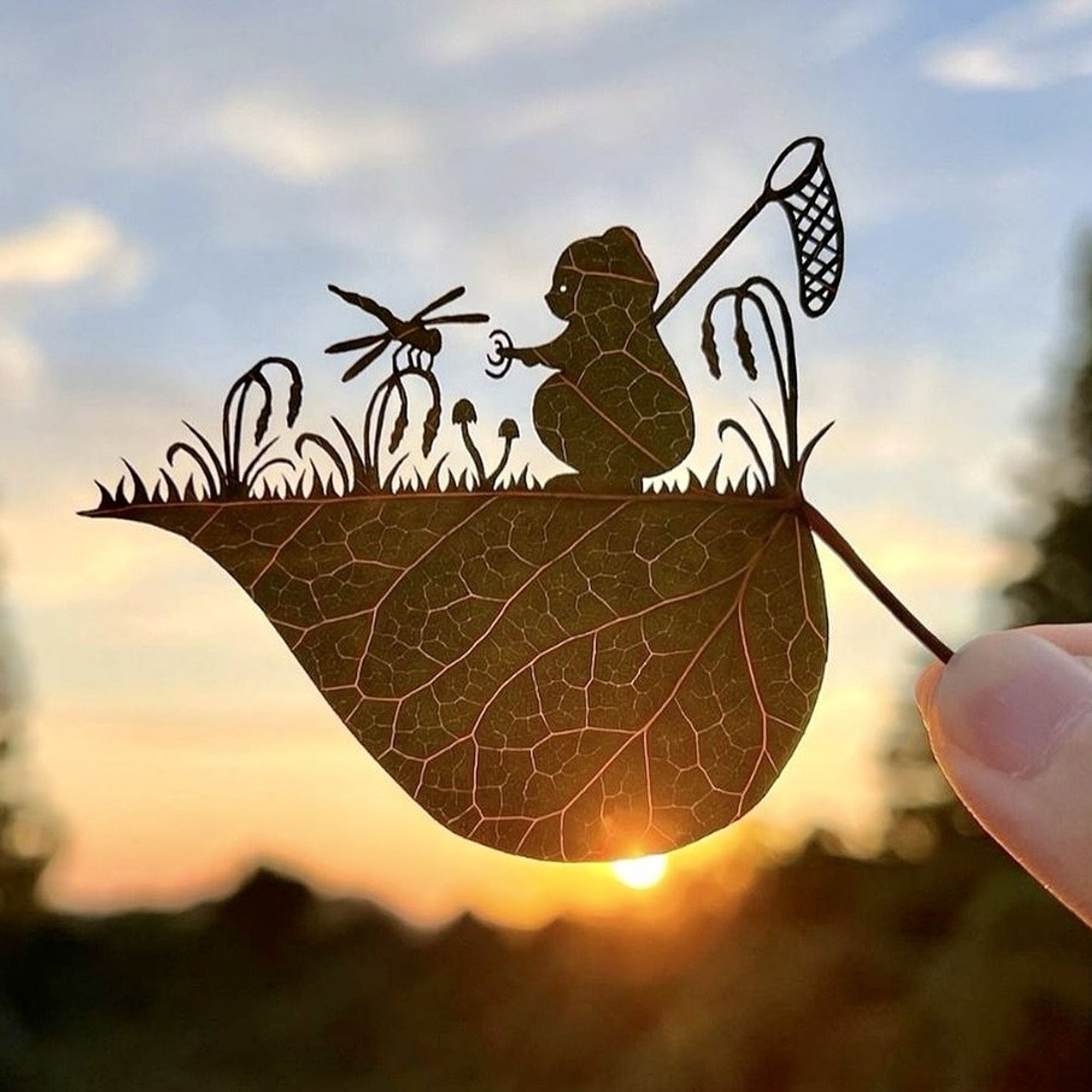 A leaf is held horizontally in front of a blue sky.  The top half is carved, the bottom half is untouched.
The scene depicts several flowers grass and two mushrooms.  On one of the flowers a drqagonfly has landed.  Next to it stands a bear (Winnie the Pooh?) with a net over his shoulder and one hand stretched out towards the dragonfly.